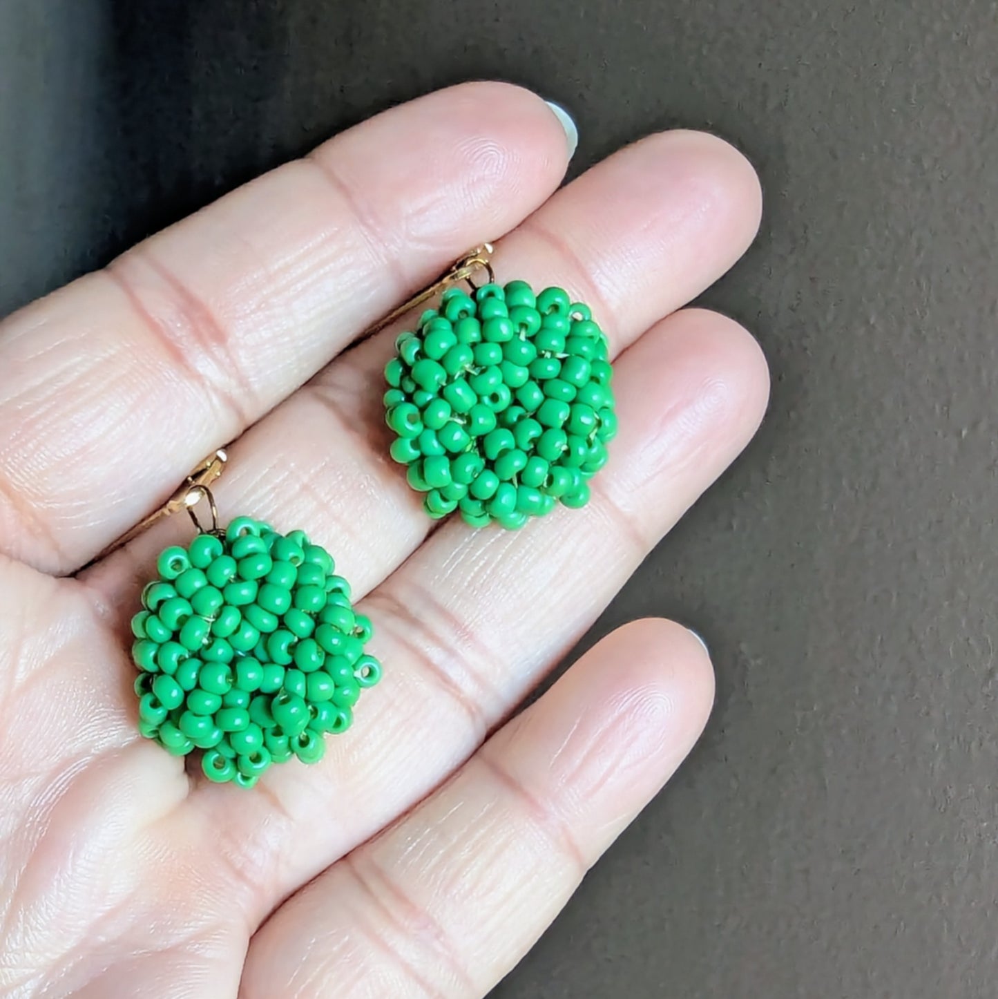 Hand holding Masi Harita earrings showing vivid green beadwork and antique bronze wire detailing.