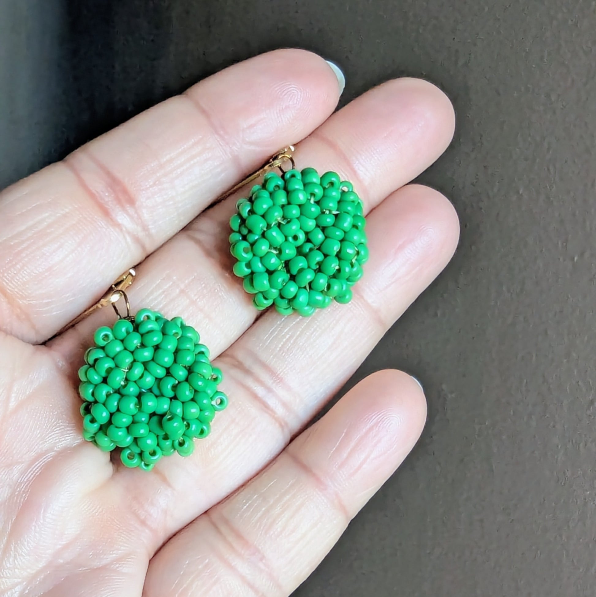 Hand holding Masi Harita earrings showing vivid green beadwork and antique bronze wire detailing.
