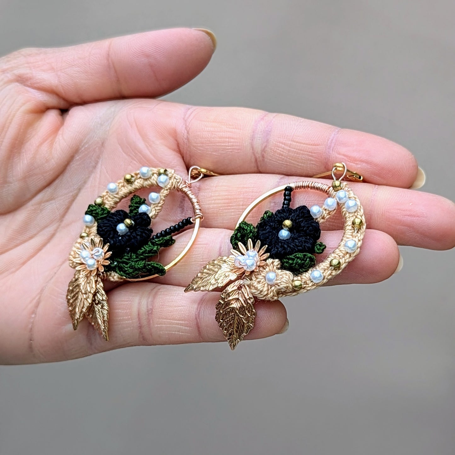 Hand presenting Mayaani Jewellery's Aloki hoops - black crochet flowers, olive leaves, pearls, and golden accents.