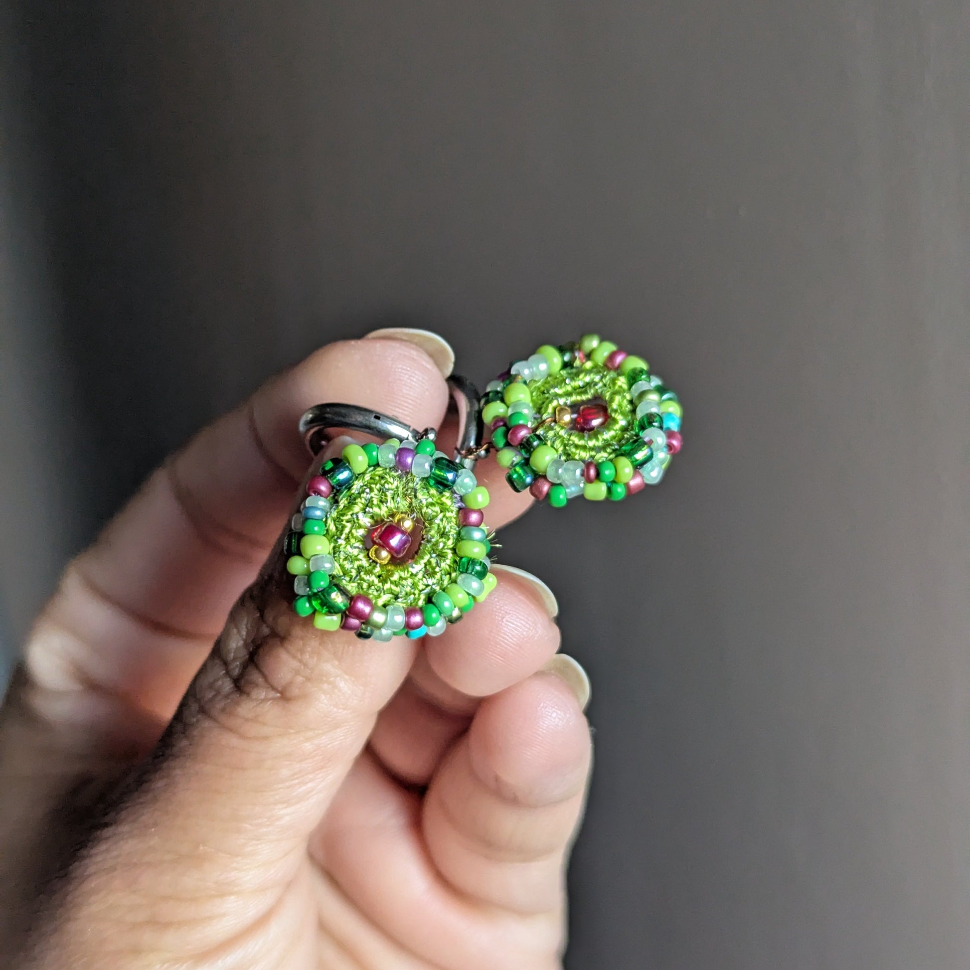 Pair of Vana artisan earrings held individually in hand, highlighting symmetry, colour variation, and the handcrafted nature of each circular bead crochet piece.