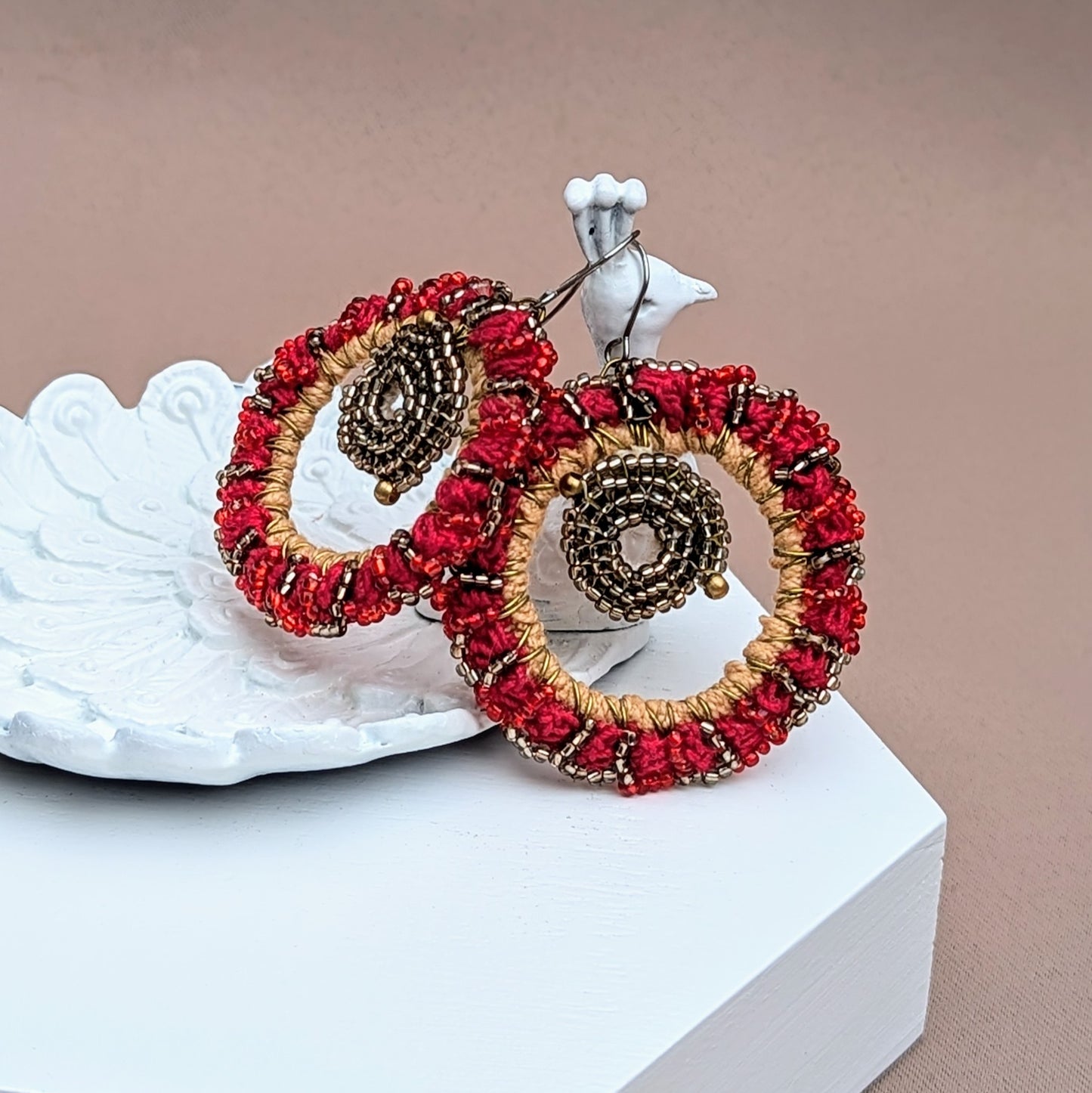 Close up view of a sustainable handmade pair of hoop earrings in radiant red and gold tones on a white peacock trinket dish exclusive to Mayaani Jewellery.
