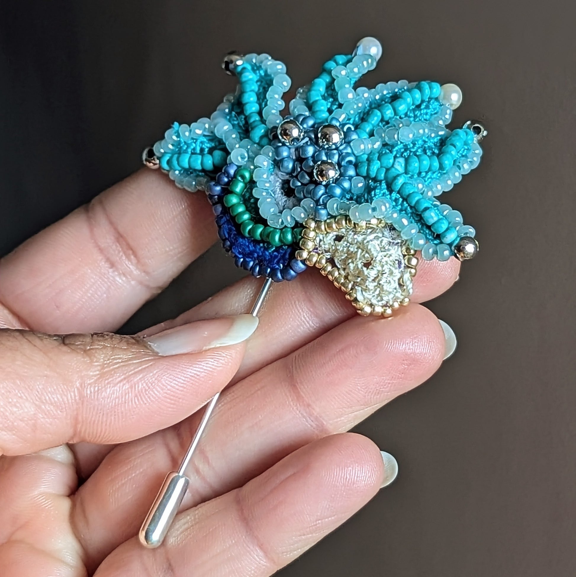 Jalika handmade turquoise beaded brooch held in hand showing sculptural seed bead petals, pearl accents and stainless steel brooch pin.