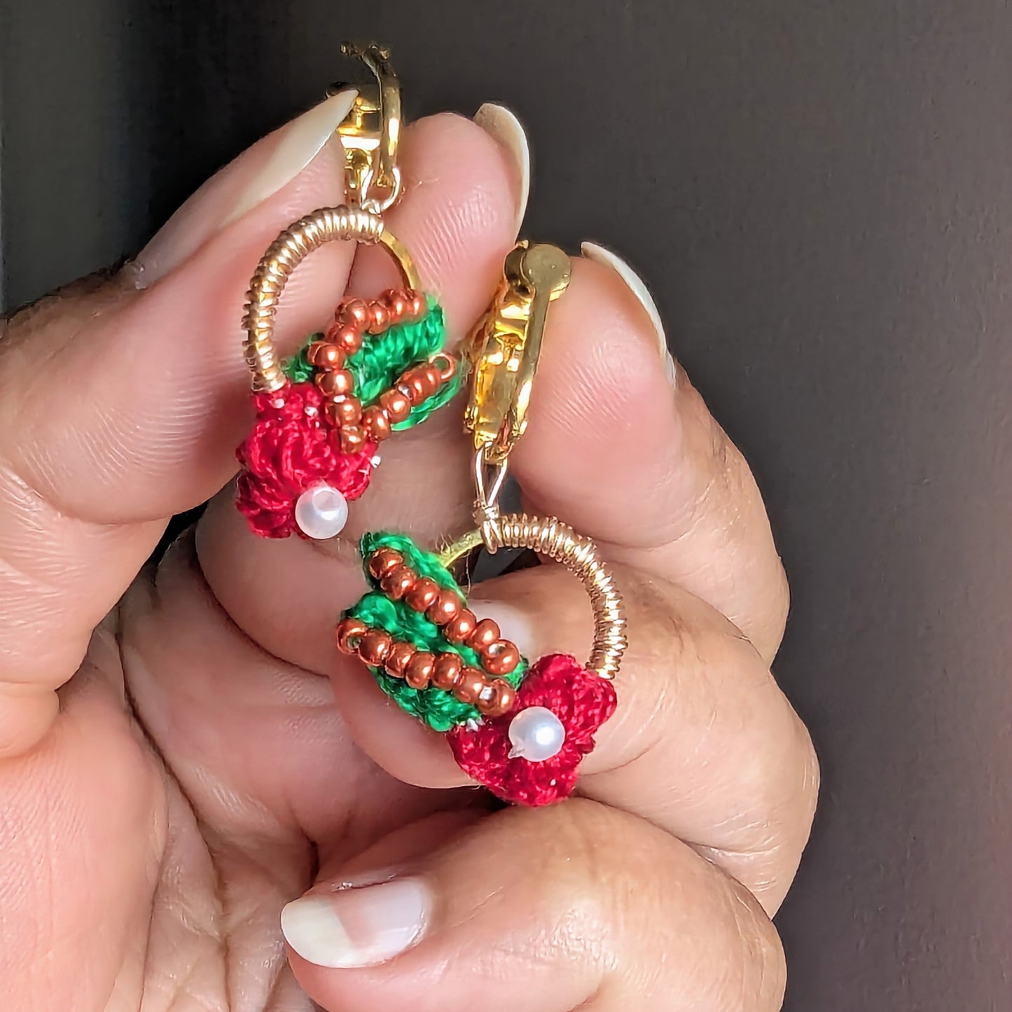 A close-up of a hand holding a pair of Kumkum handmade beaded clip-on earrings. The earrings feature gold stainless steel hoops and clip-on screw back clasps decorated with red and green seed beads, small brown beads, and white pearl-like accents.