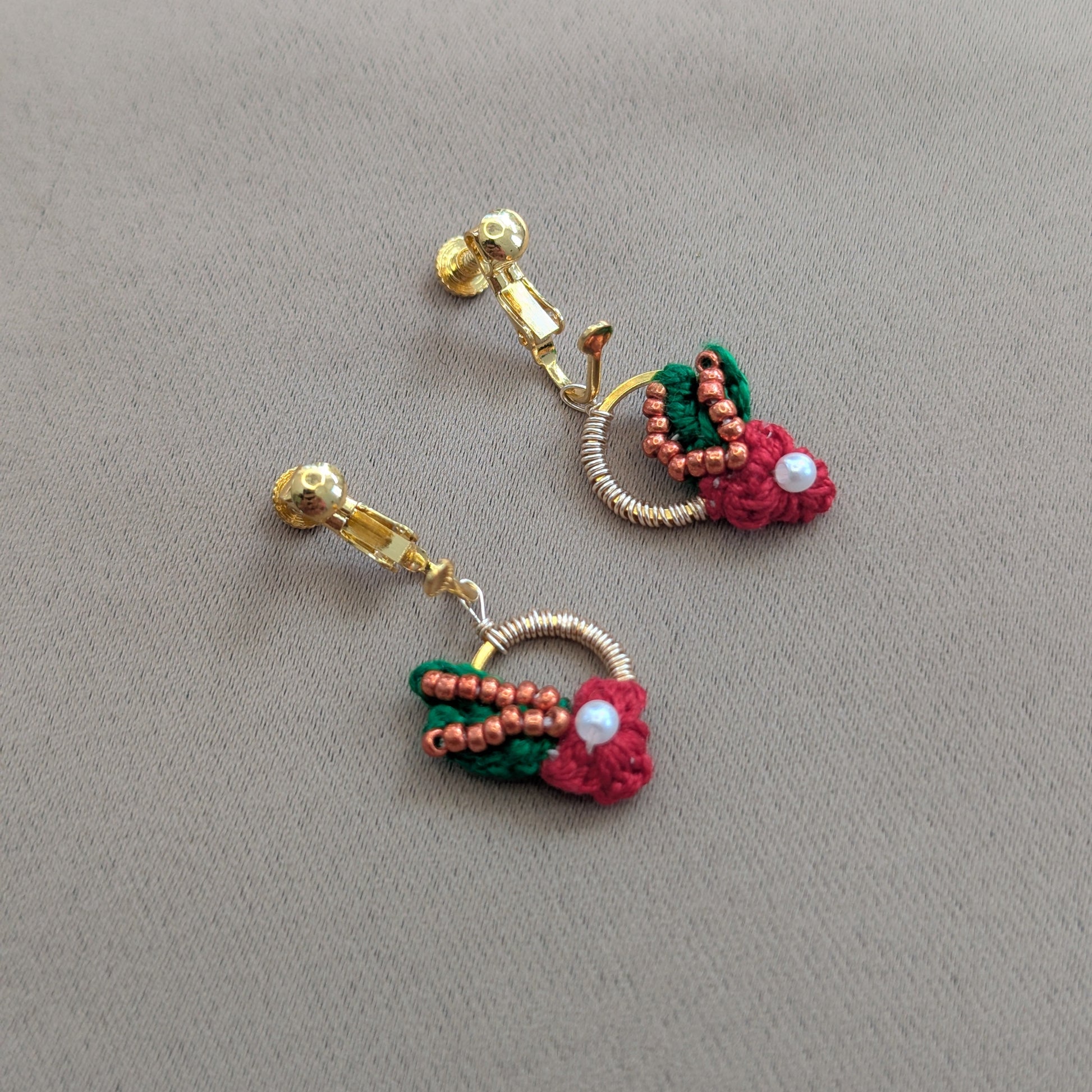 Close up view of a pair of small dangle drop style lightweight comfortable red, copper, gold and green clip-on earrings called Kumkum exclusively available at Mayaani Jewellery UK.