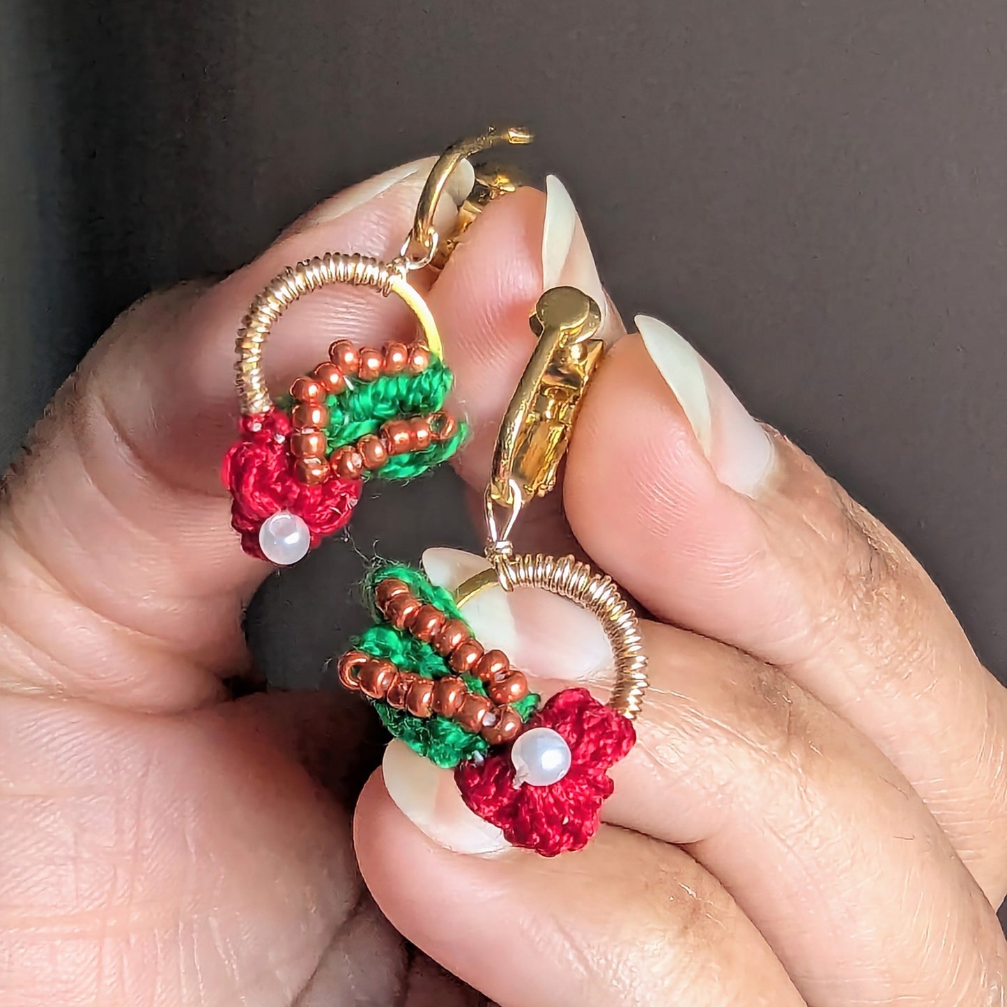 Close up of a person holding a delicate pair of sustainably handmade Kumkum earrings in red floral motifs with green, copper, pearl and rose gold accents exclusively created by Mayaani Jewellery UK.