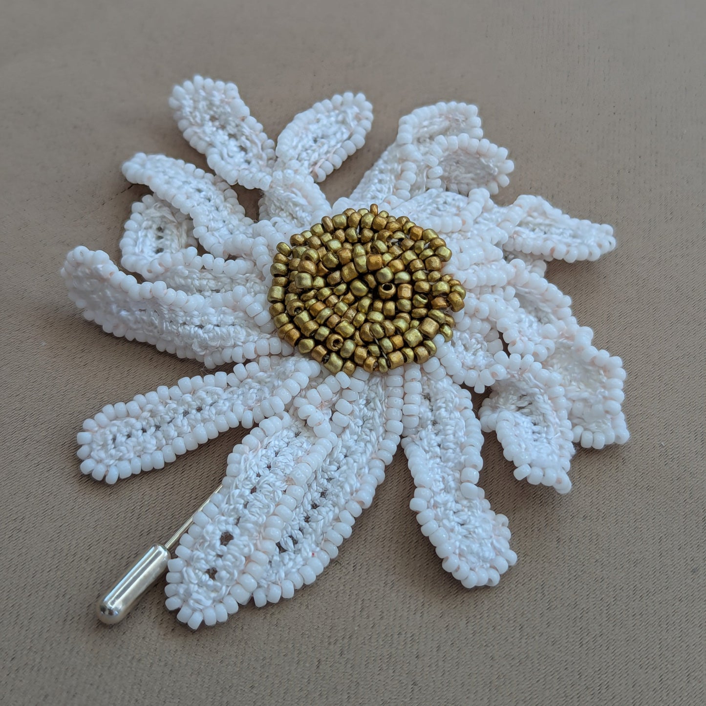 Flat lay of a one of a kind daisy brooch lapel hat pin on soft grey backdrop featuring hand crochet petals edged in white seed beads meeting at a gold seed bead centre.