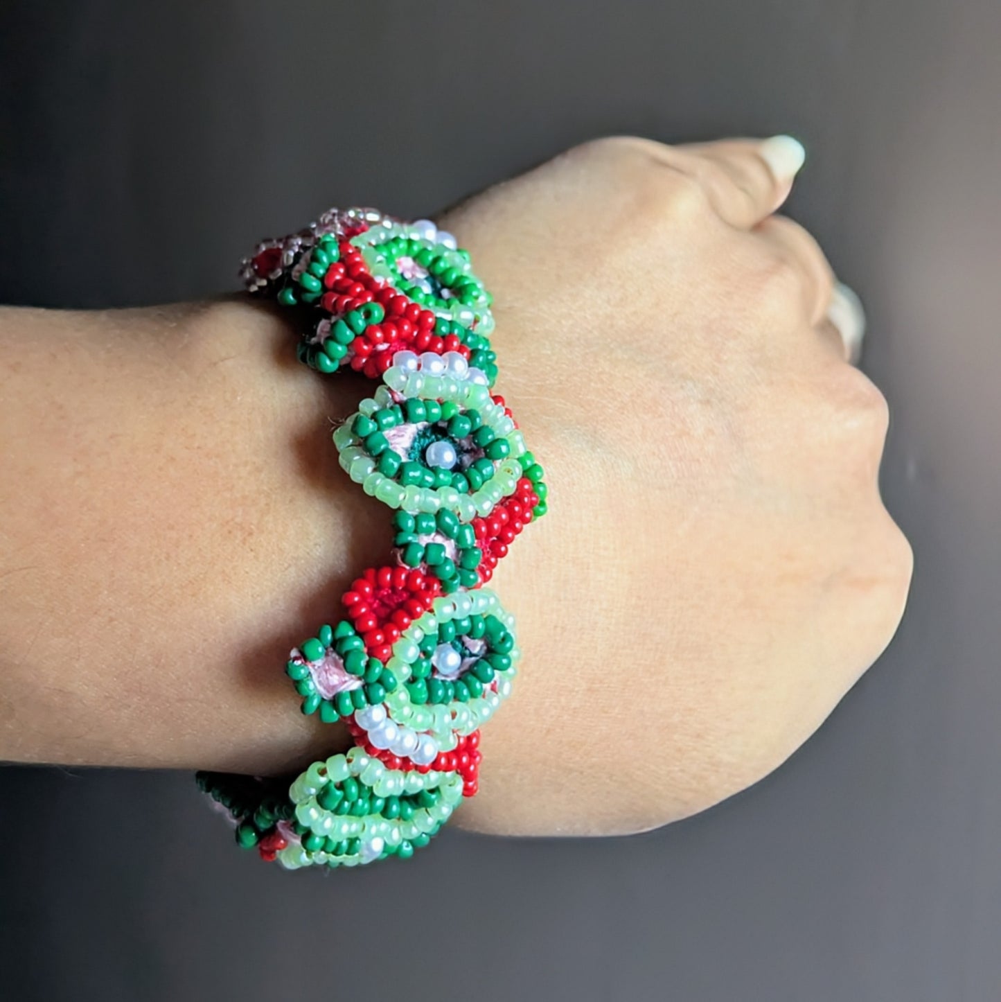 A red and green bead and crochet woven intricate hand bracelet modelled on a wrist against a warm grey backdrop.