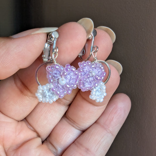 Handmade beaded Malti clip-on hoop earrings held in a hand. The earrings feature light purple flower-shaped beads with white pearl-like accents and silver-toned metal hoops and ear clips.