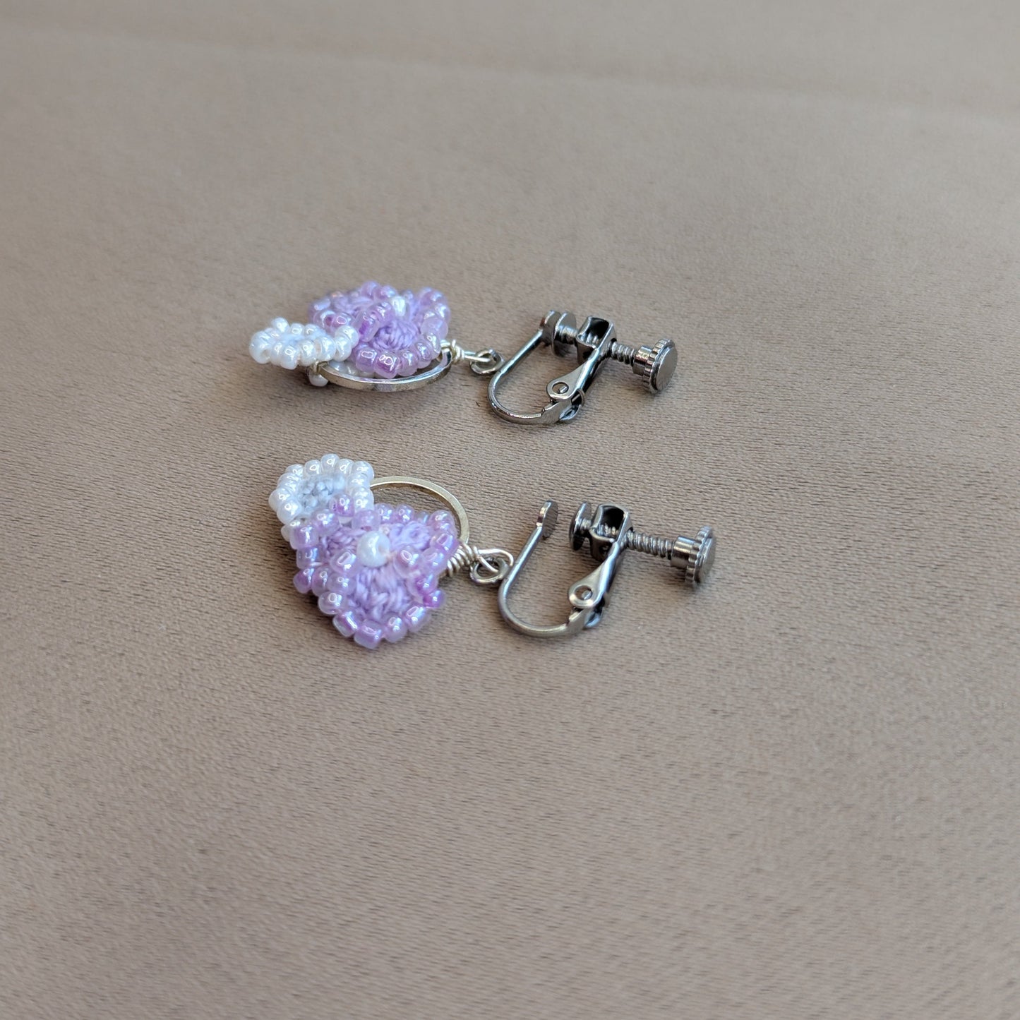 A pair of Malti clip-on earrings handmade  using beaded hoops featuring light purple and white seed beads, attached to silver-toned metal hoops with screw-back clasps, displayed on a textured beige surface.