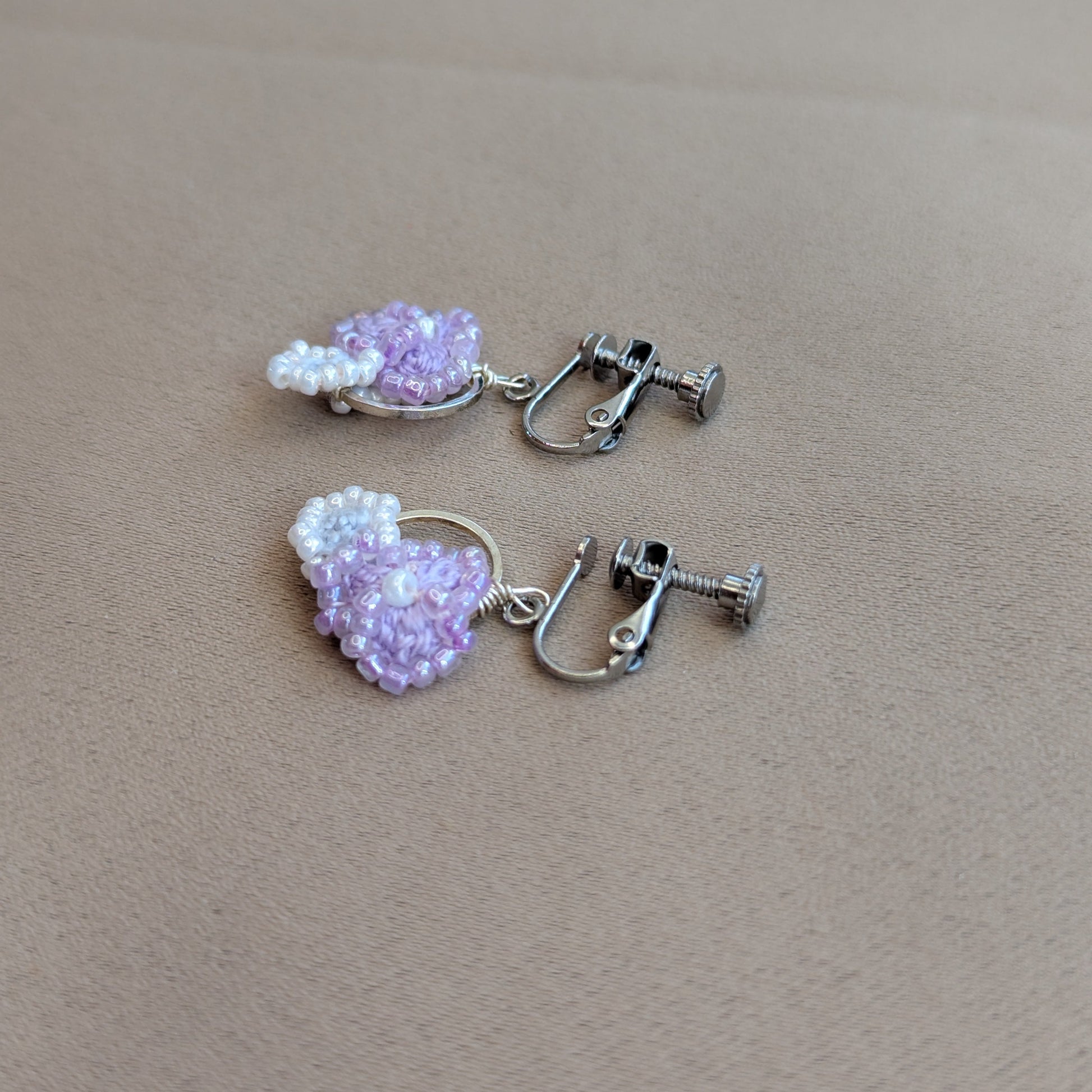 A pair of Malti clip-on earrings handmade  using beaded hoops featuring light purple and white seed beads, attached to silver-toned metal hoops with screw-back clasps, displayed on a textured beige surface.
