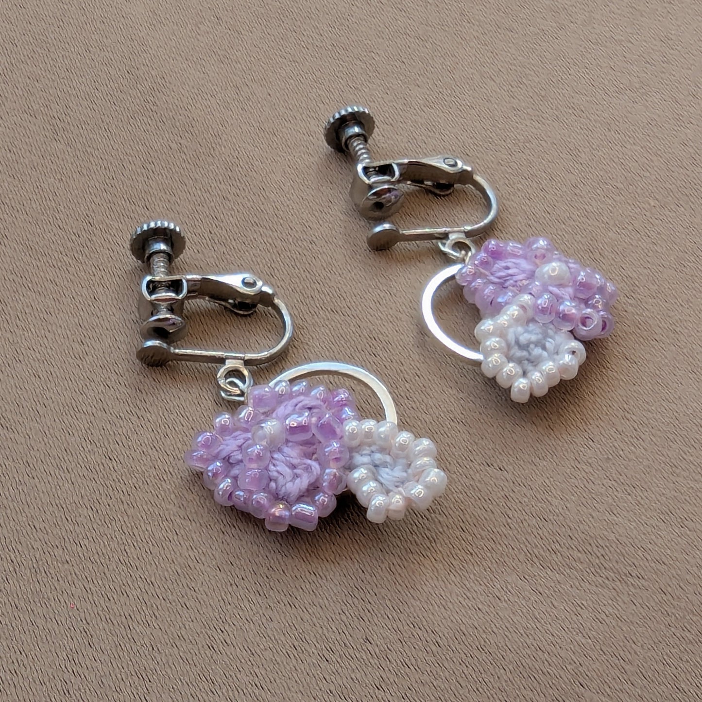 Malti handmade lilac, silver, white dangle drop clip-on earrings featuring crochet and beaded floral elements on a silver hoop displayed on a neutral fabric backdrop.
