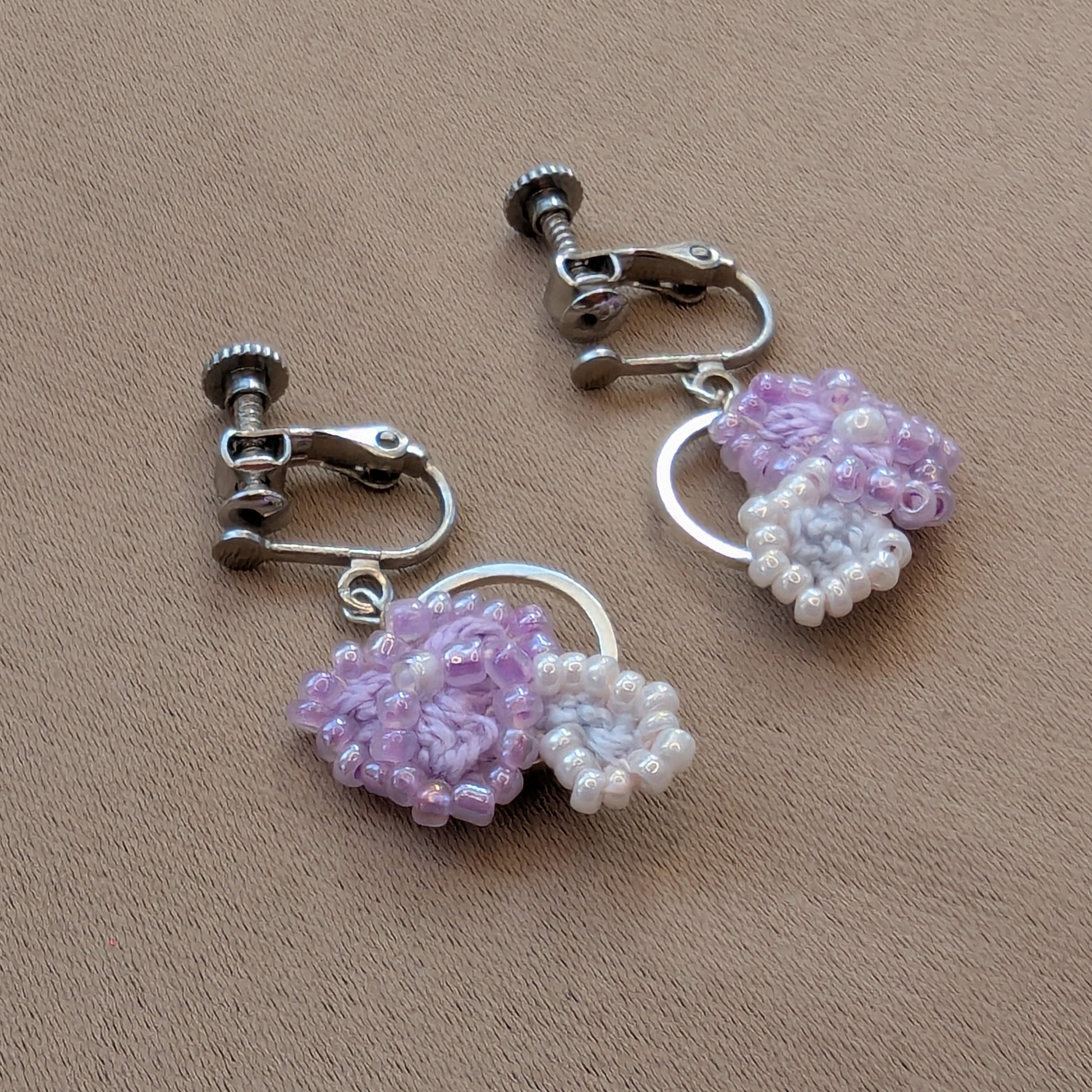 Malti handmade lilac, silver, white dangle drop clip-on earrings featuring crochet and beaded floral elements on a silver hoop displayed on a neutral fabric backdrop.