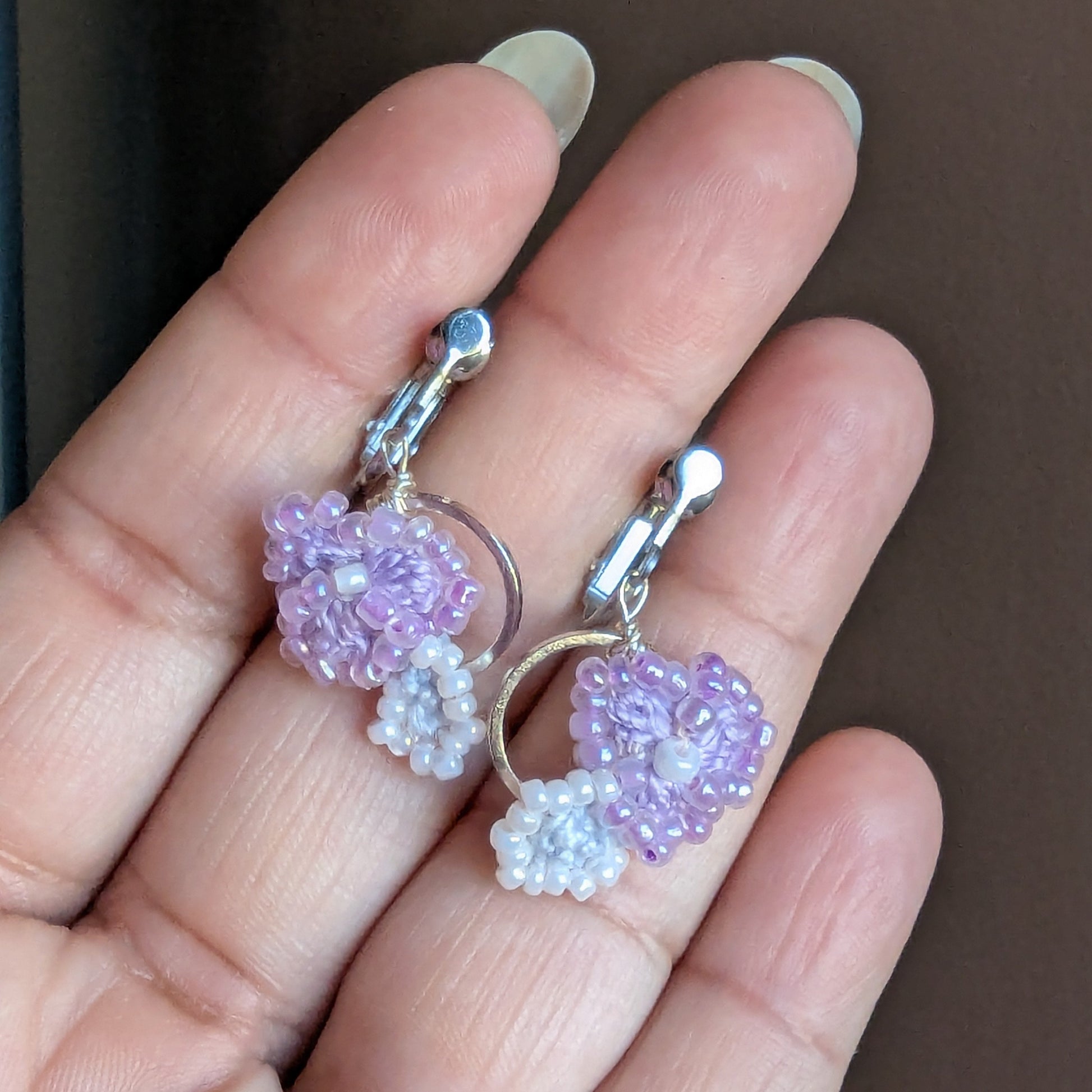 Hand holding the Malti artisan made pair of clip-on earrings featuring a silver stainless steel hoop with delicate crochet and beadwork floral elements unique to Mayaani Jewellery UK.