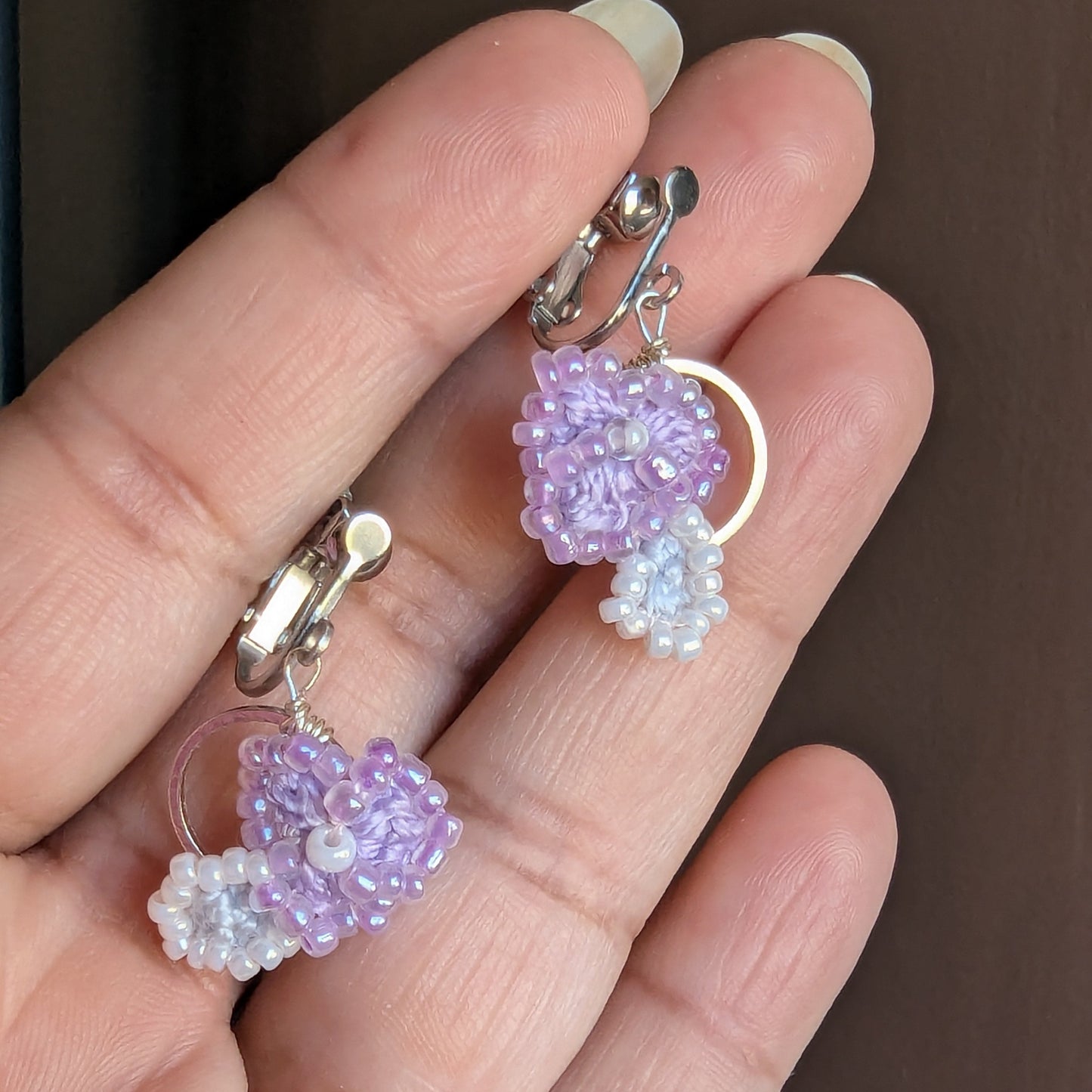 Malti clip-on earrings featuring a circular silver hoop, lilac and white crochet botanical details and fine seed beads held gently between fingers against a grey backdrop.