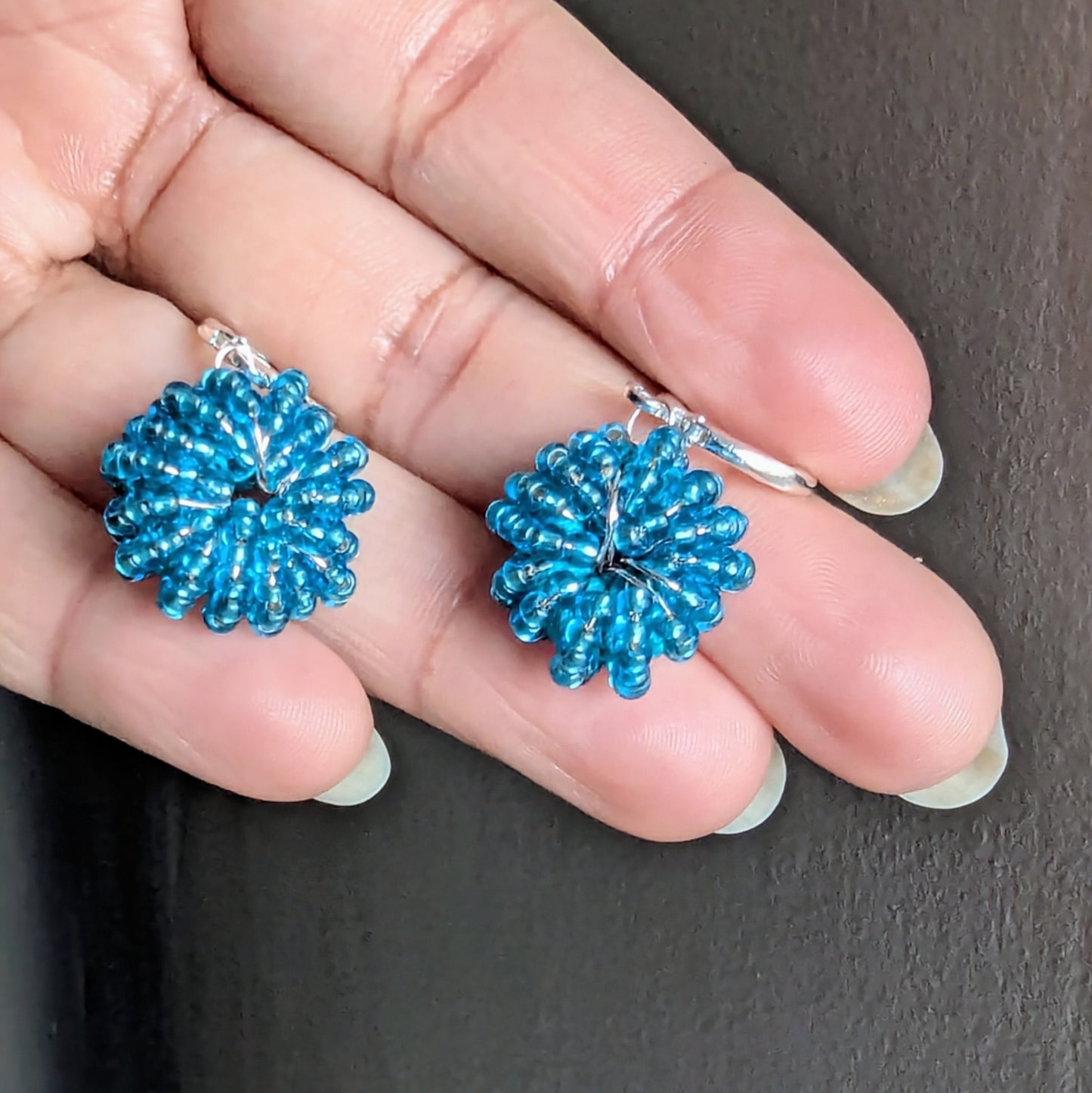 Small round blue handmade earrings held between fingers, shimmering with silver-lined beads and rose gold accents.