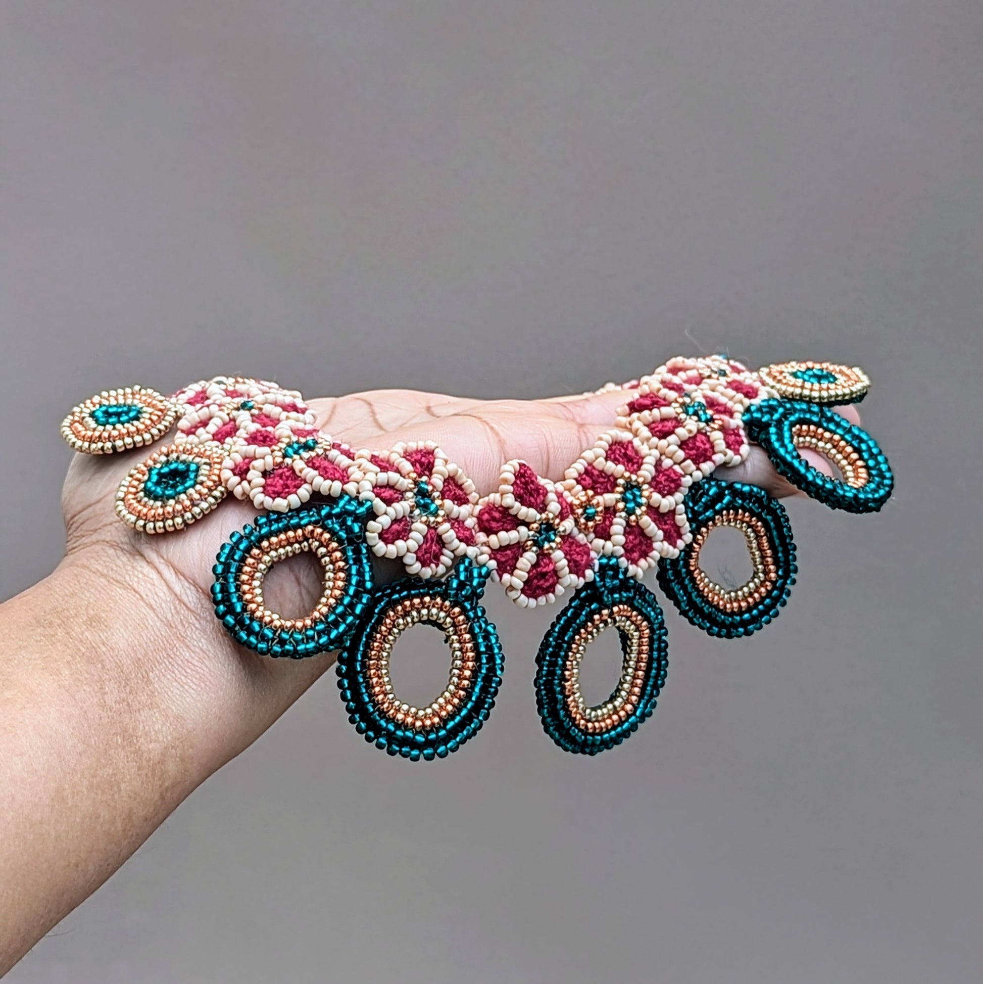 Hand holding a colourful red, emerald green, peach and gold beaded designer bridal neckpiece designed and crafted for zero waste business Mayaani Jewellery UK.
