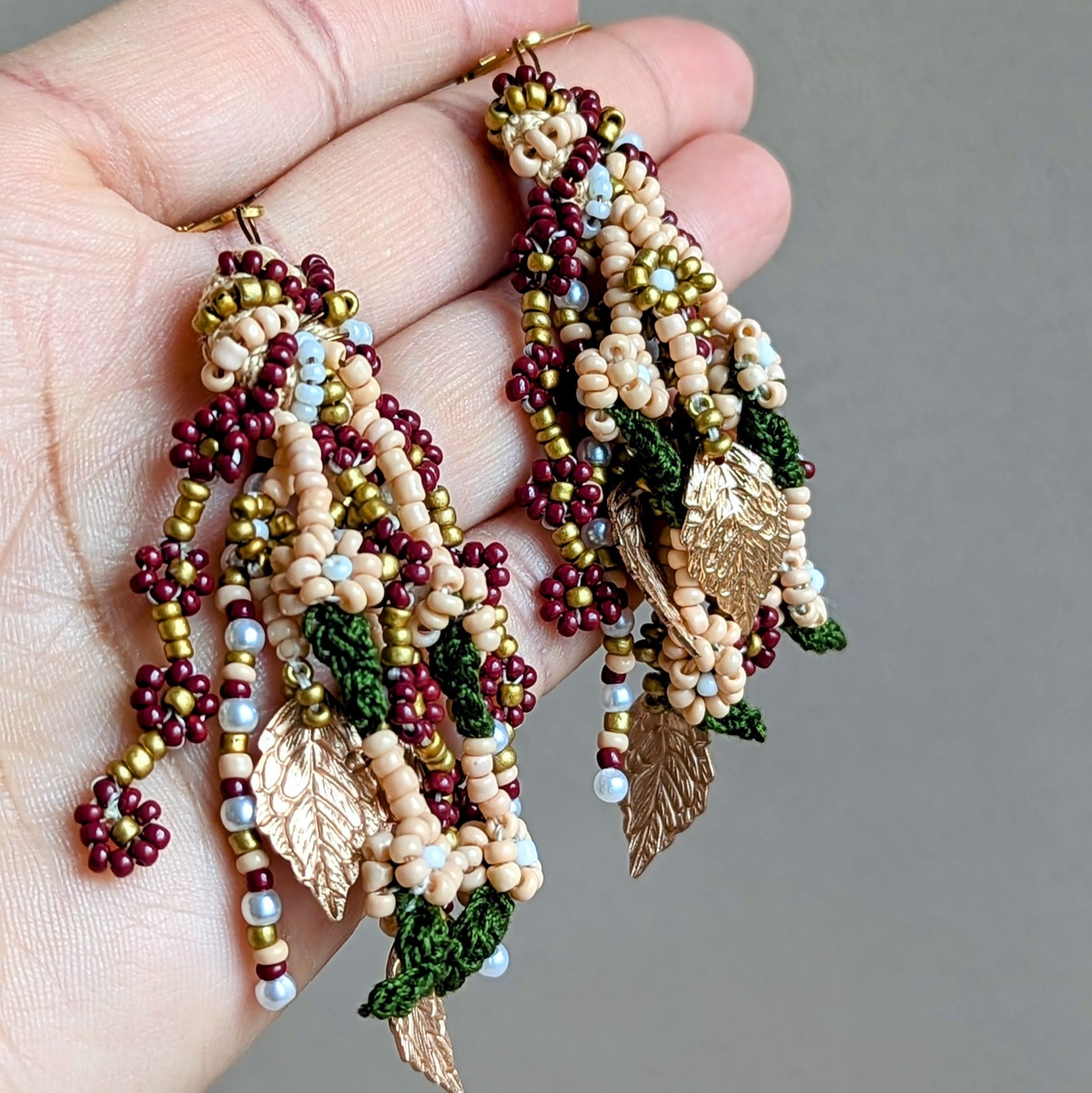 Elegant handcrafted Indian earrings photographed in the palm of a hand. Designed for weddings and special occasions, these multi-strand Mayaani earrings feature red, green, gold and pearl seed beads with gold leaf charms.