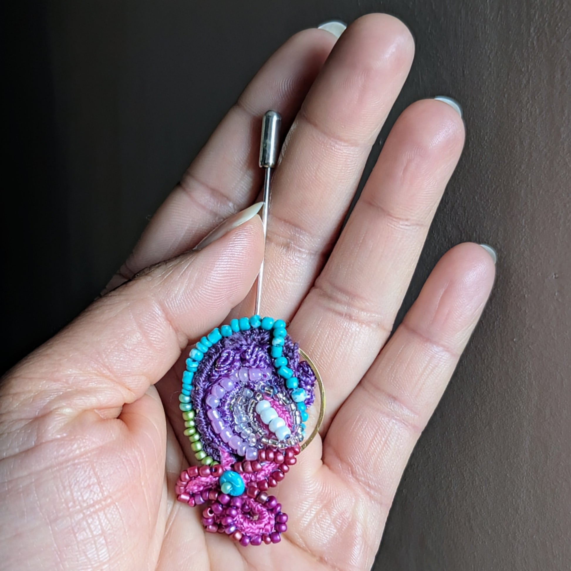 Navira sustainable statement brooch resting in palm of hand to show scale, featuring layered purple and turquoise bead embroidery with gold hoop detail.