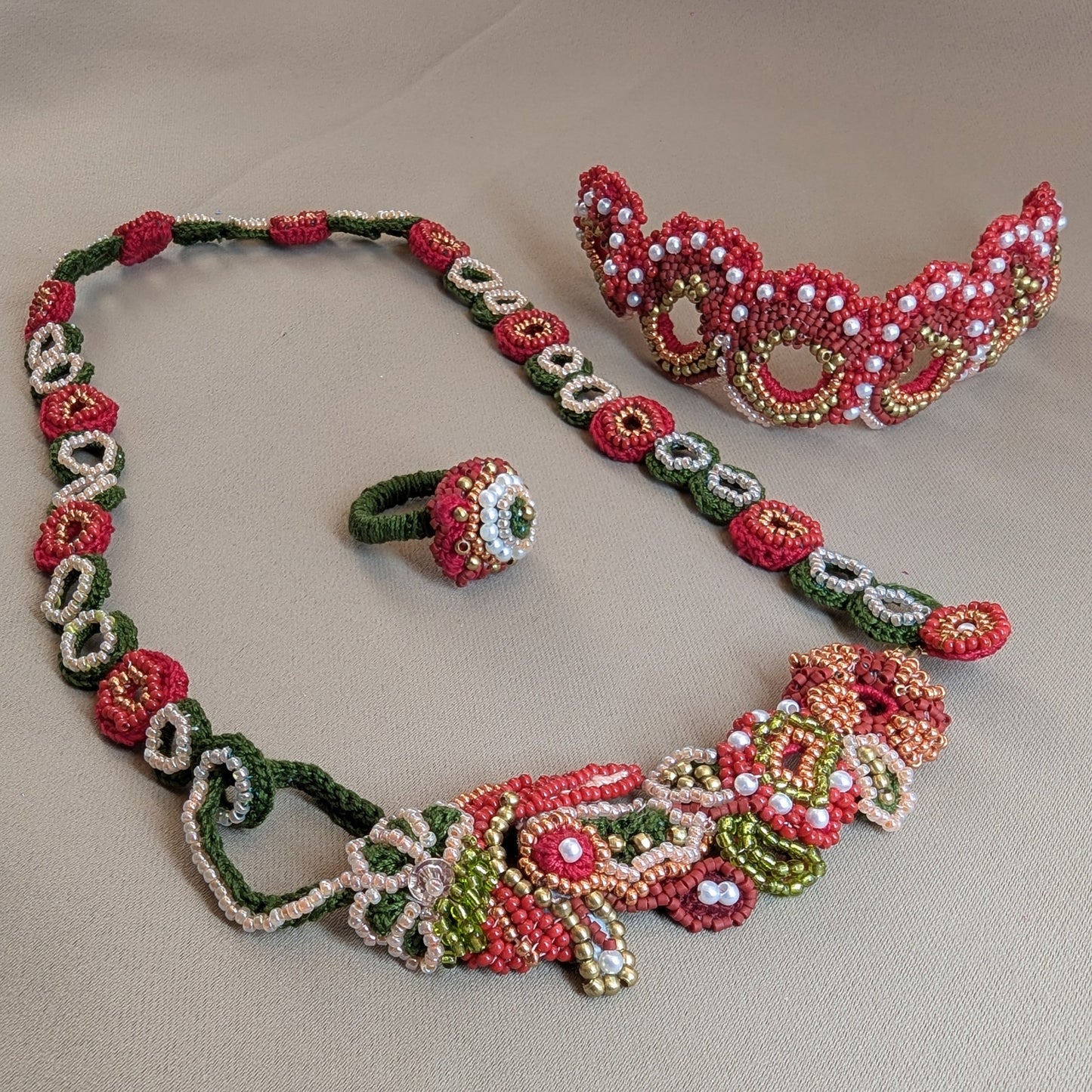 Shreya 5-in-1 artisan beaded jewellery set featuring a necklace, a ring, and a bracelet, heavily beaded in red, green, gold and soft peach beads in floral and geometric patterns, displayed on a neutral beige background. 