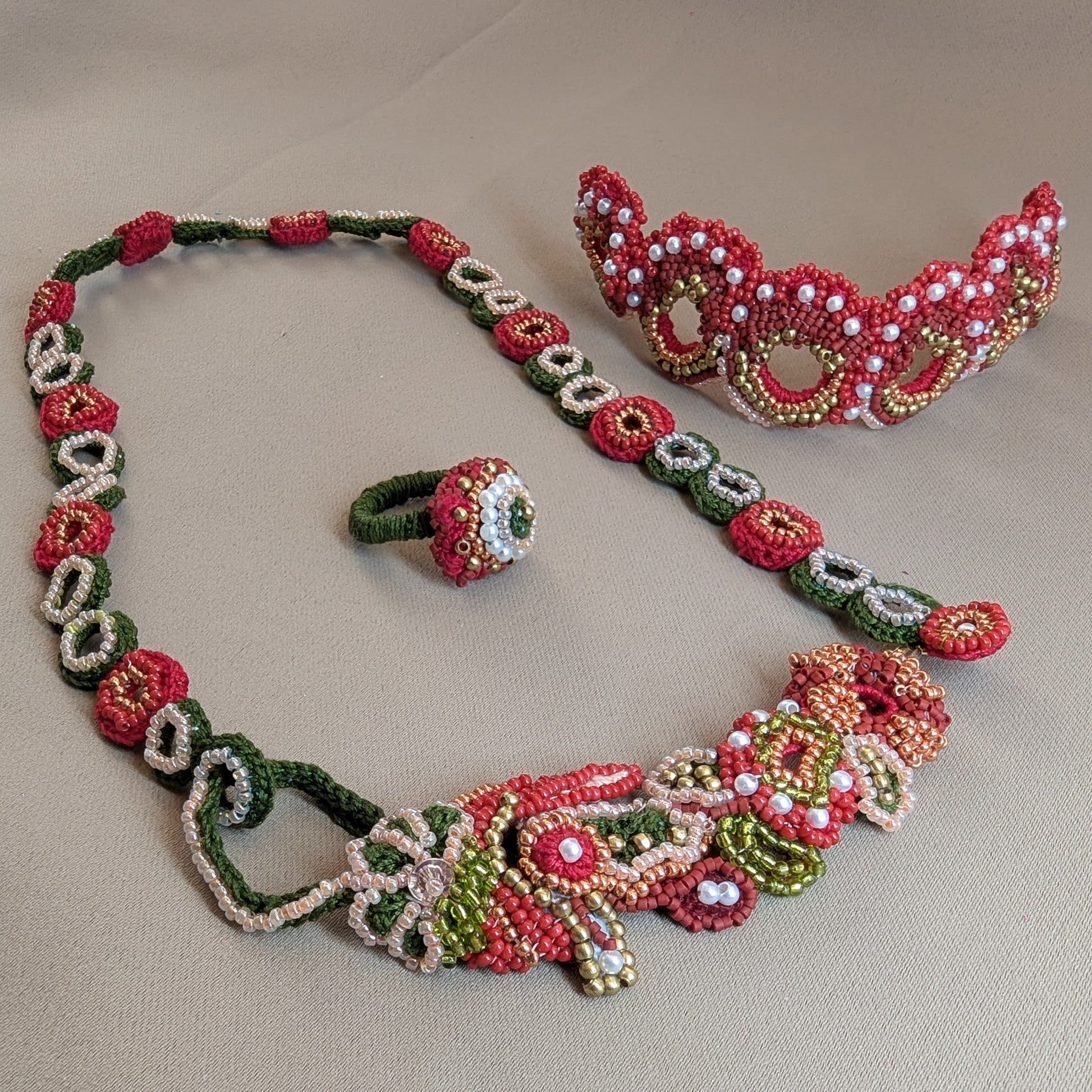 Shreya 5-in-1 artisan beaded jewellery set featuring a necklace, a ring, and a bracelet, heavily beaded in red, green, gold and soft peach beads in floral and geometric patterns, displayed on a neutral beige background. 