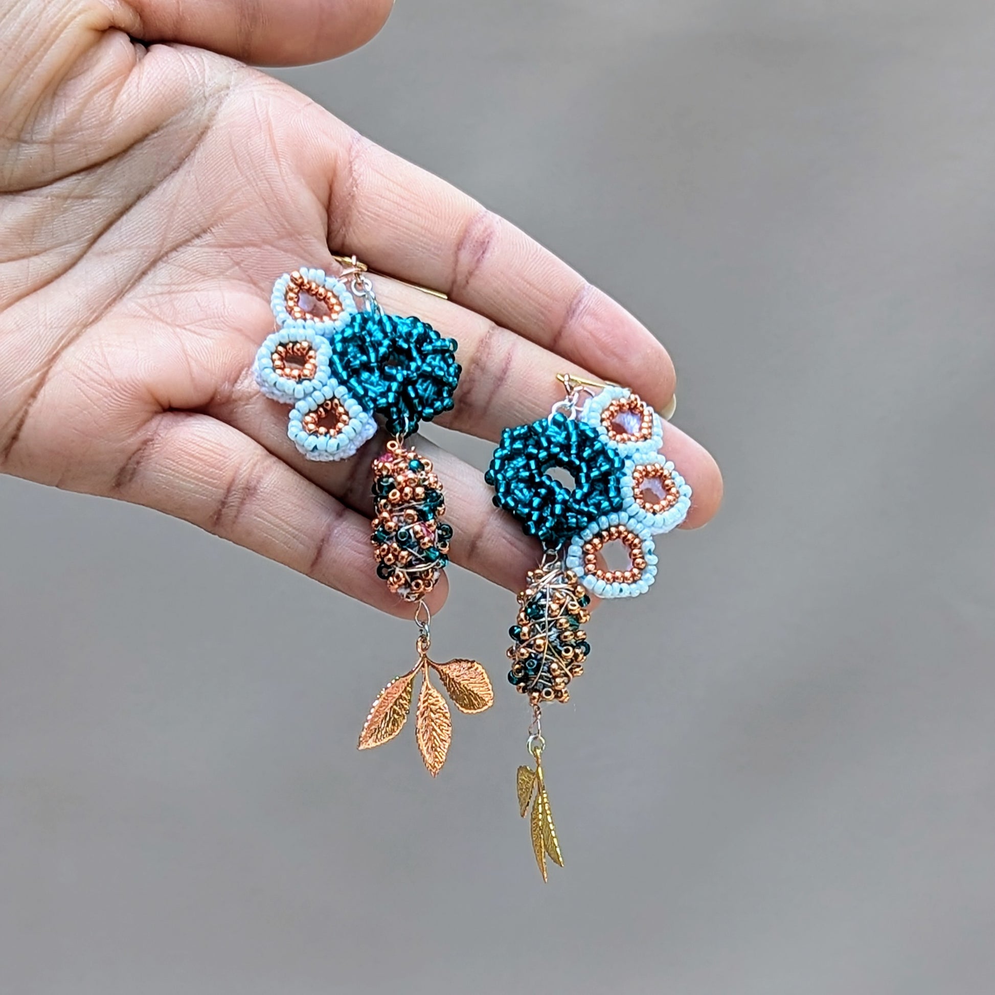 Hand holding an asymmetric scrap bead pair of Mayaani Jewellery earrings handcrafted in the UK using high quality glass seed beads in white, emerald green and rose gold alongside beads created using scraps from the jewellery making process.