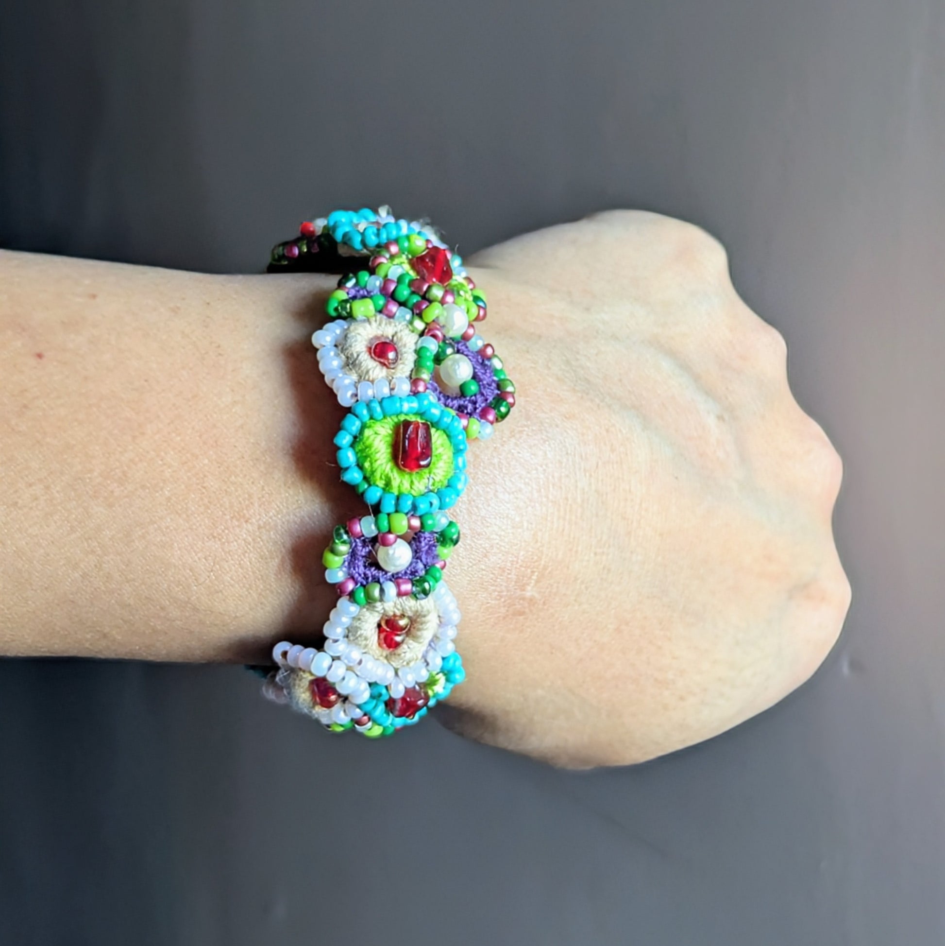 Handcrafted beaded bracelet worn on the wrist, featuring circular crochet motifs with green, turquoise, pearl, and red new and vintage glass seed beads, inspired by a blooming garden and handmade in the UK.