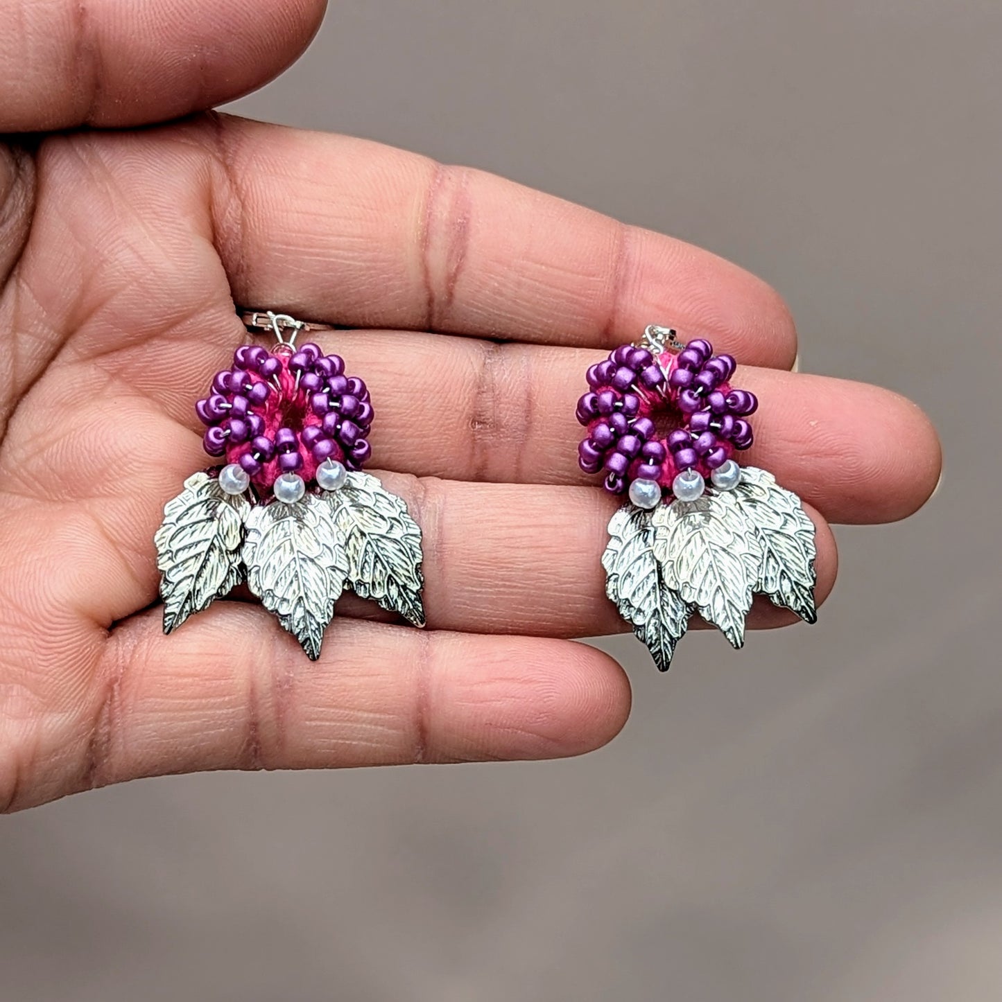 Hand holding Vayupatra handmade silver leaf earrings featuring purple seed beads and sterling silver hooks.