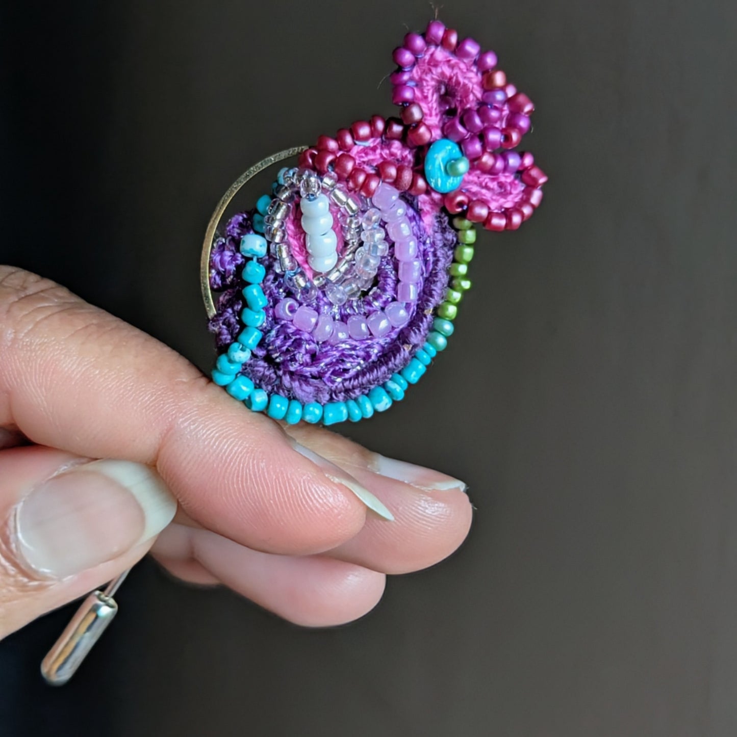 Vertical composition of Navira one of a kind artisan brooch held against neutral wall background highlighting colourful beadwork and gold hoop accent.
