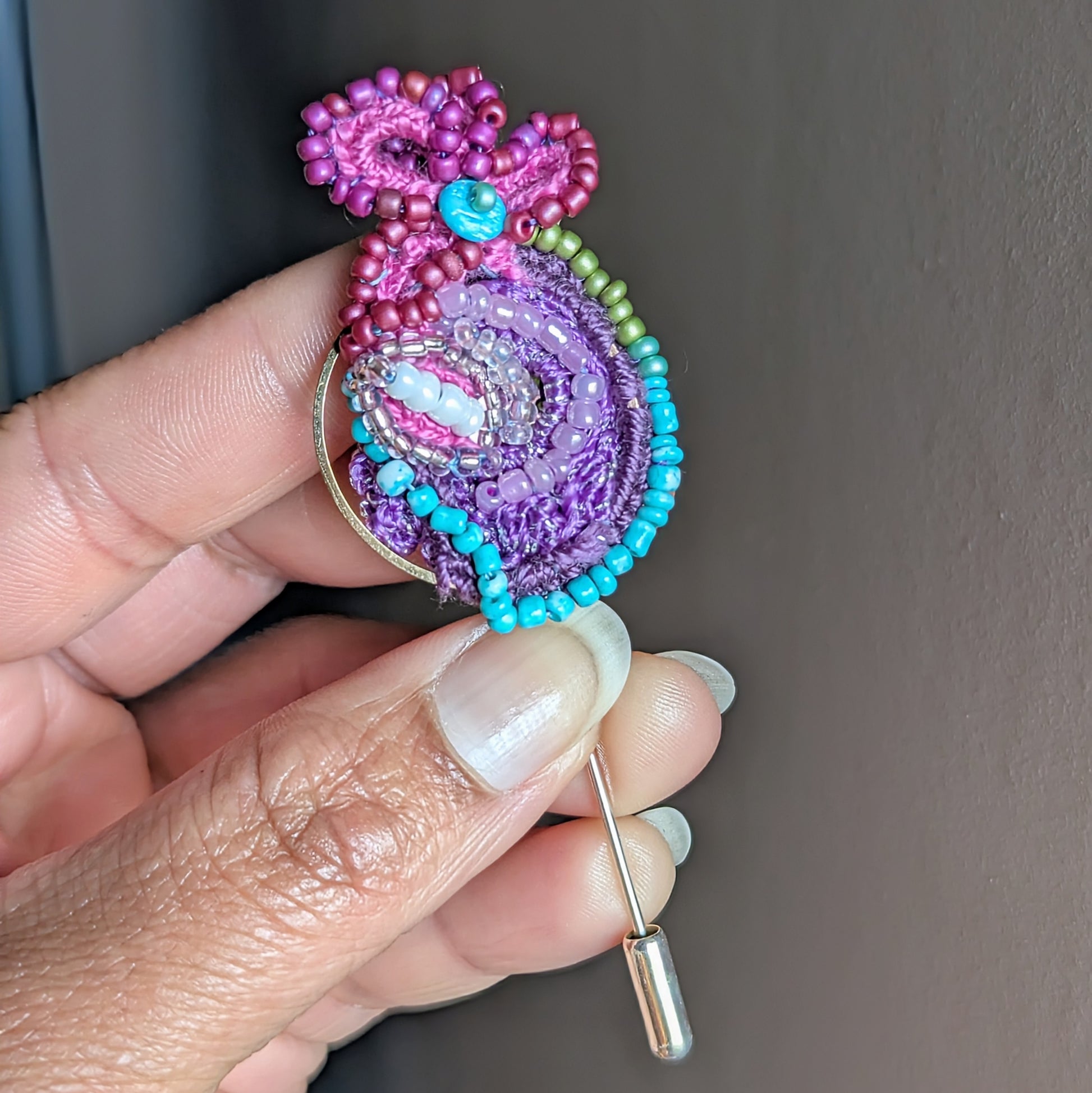 Navira one of a kind handmade beaded brooch held in hand, featuring layered purple, turquoise and pink glass seed beads with gold hoop detail and stainless steel pin fastening.