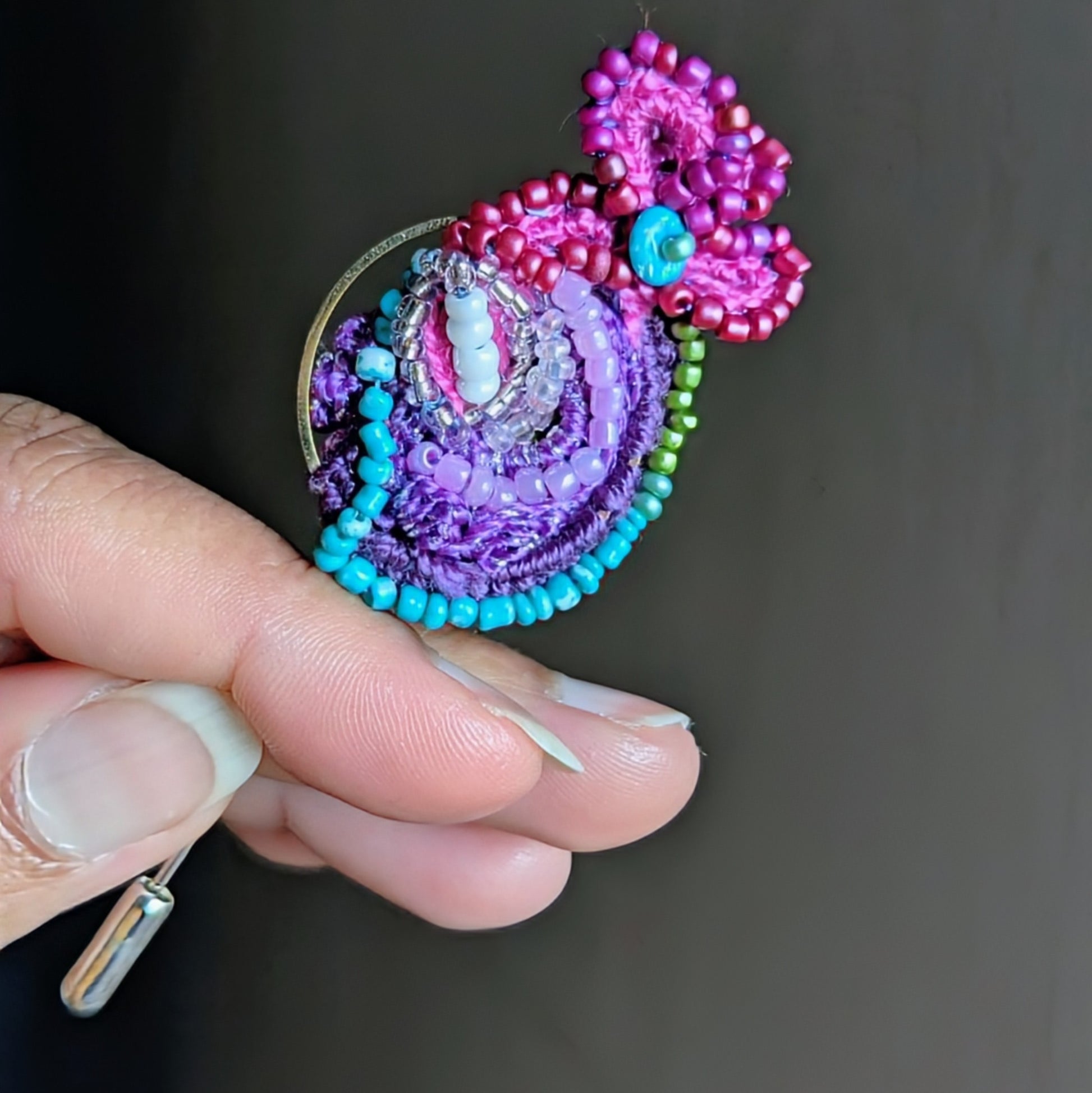 Handheld view of Navira one of a kind beaded statement brooch in purple, turquoise  and pink tones with gold hoop detail, handcrafted using glass seed beads and Egyptian cotton threads.