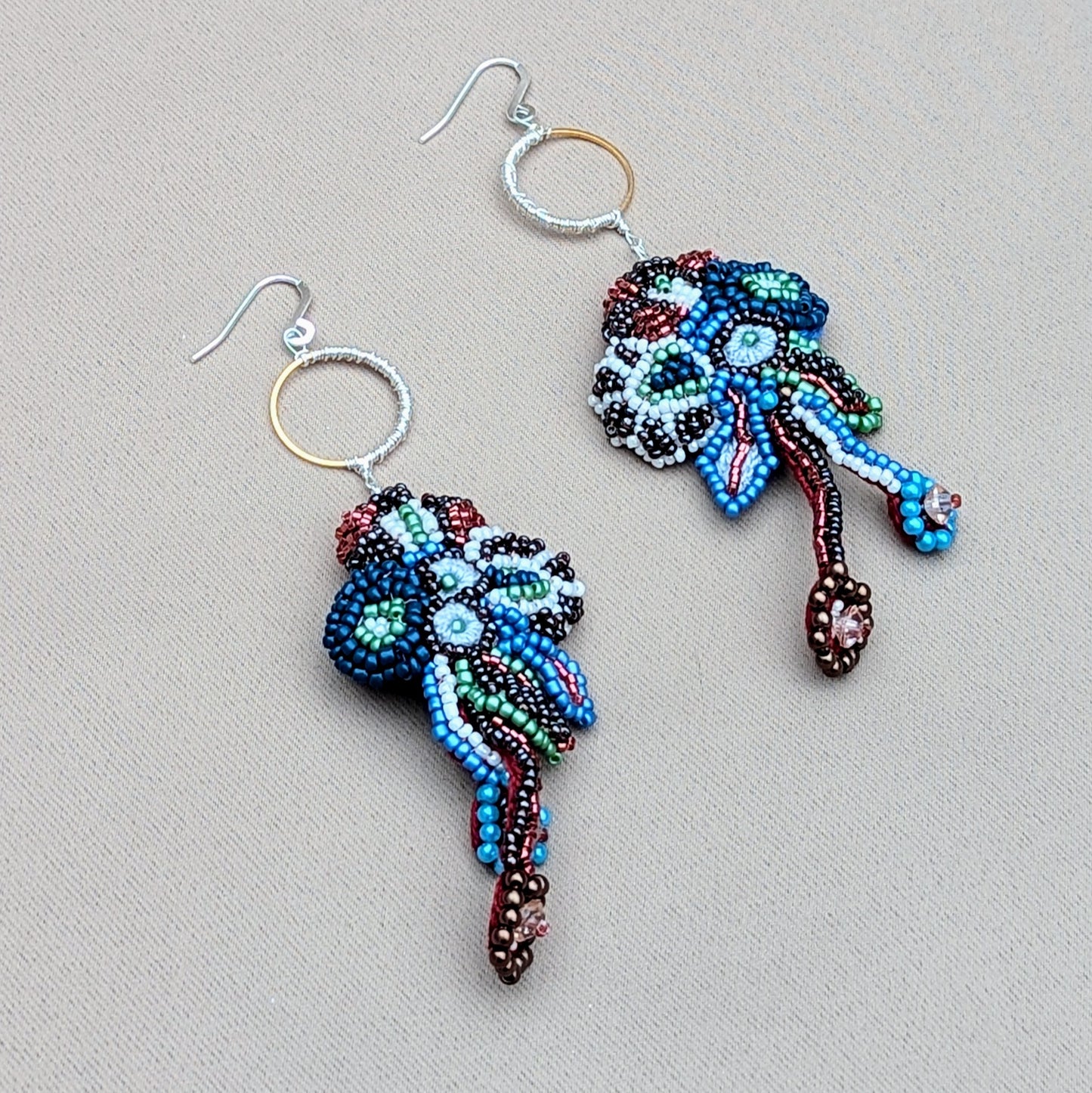 Flat lay view of one-of-a-kind Neelara statement earrings handcrafted with red, blue and white glass seed beads, featuring flowing beadwork, gold stainless steel hoops and sculptural movement, photographed on a neutral background.
