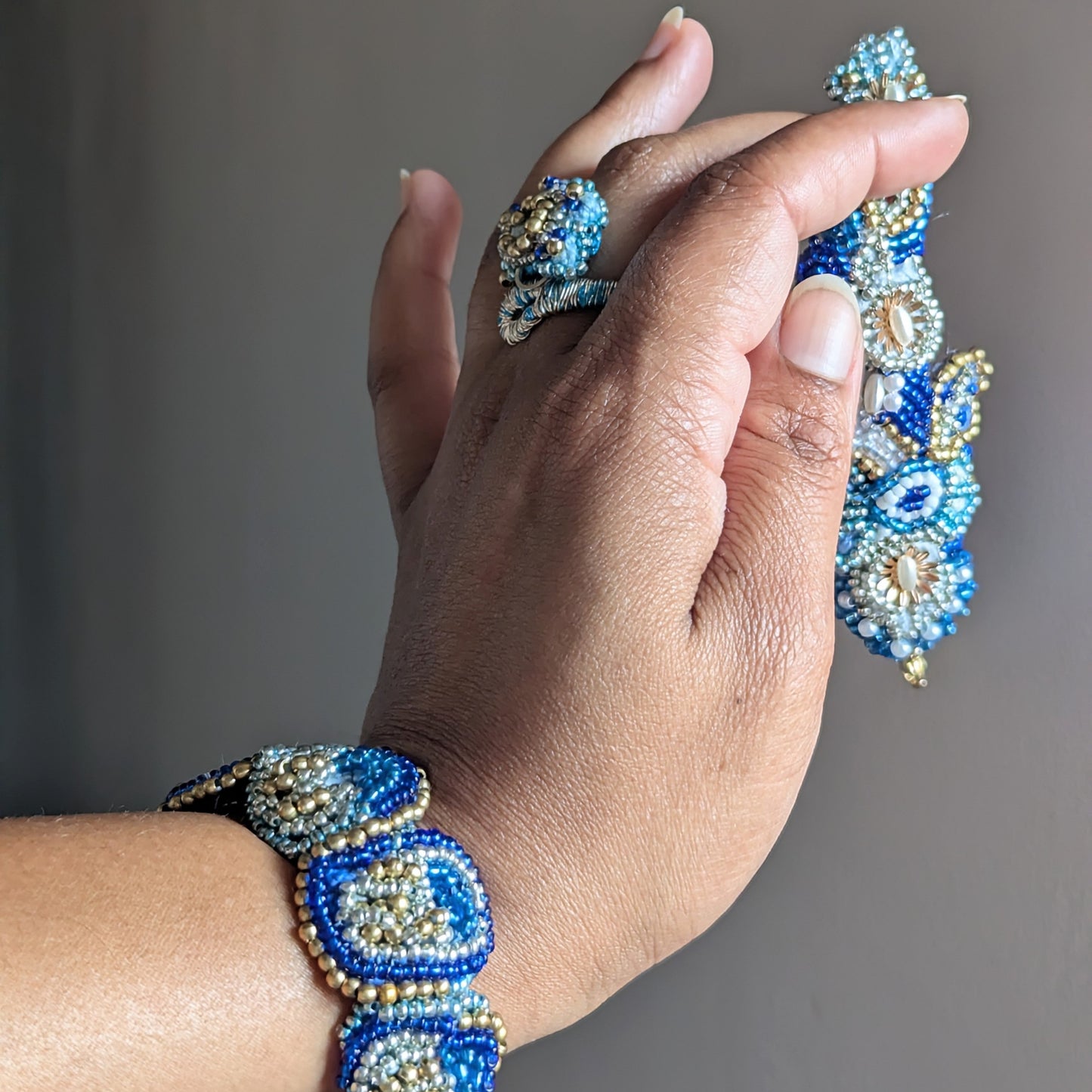 A close-up of a hand wearing the Nira 5-in-1 wrist bracelet and ring elements in blue, white and gold holding the central bracelet element against a plain grey background.