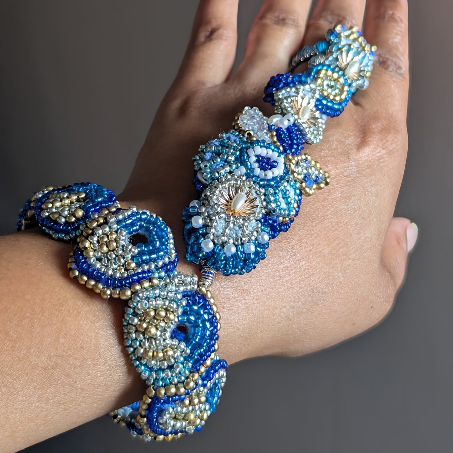A close-up of the Nira 5-in-1 Mayaani hand and wrist bracelet hand beaded with  Japanese Miyuki seed beads in blue and white, and gold solid brass beads against a neutral grey background.