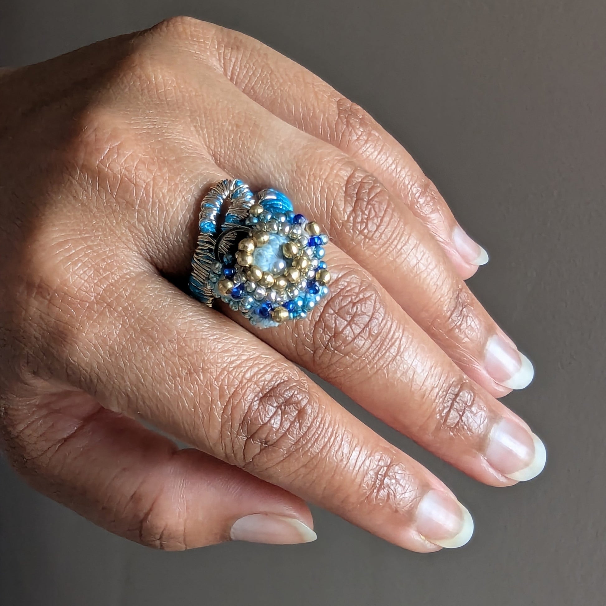 A close-up of a hand wearing the Nira 5-in-1 large, ornate ring element with blue beads, silver-toned metal, and a central pale blue stone, set against a plain grey background.