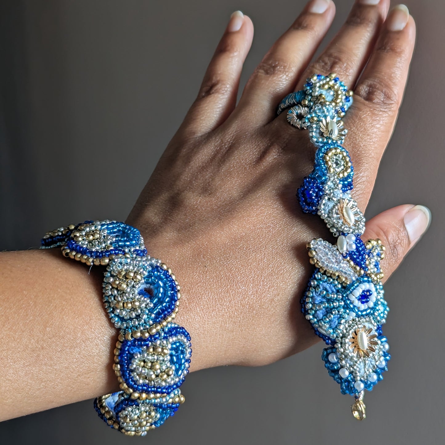 A close-up of a hand wearing the Nira 5-in-1 beaded bracelet and ring elements. The jewellery features intricate blue, white, and gold seed bead artisan craftsmanship.