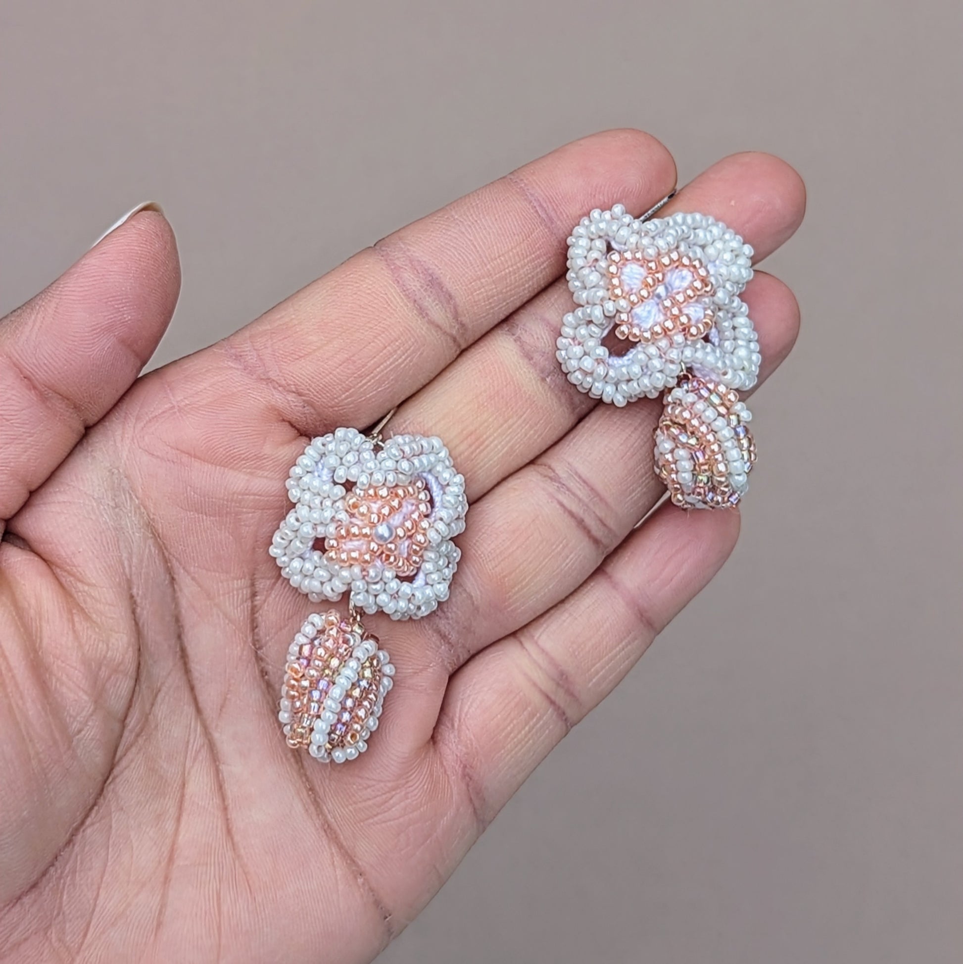 Eco-friendly earrings in a soft bridal palette of peach and white, displayed against a neutral linen backdrop. The design features seed bead florals, upcycled scrap beads, and 25 silver lever-back hooks.