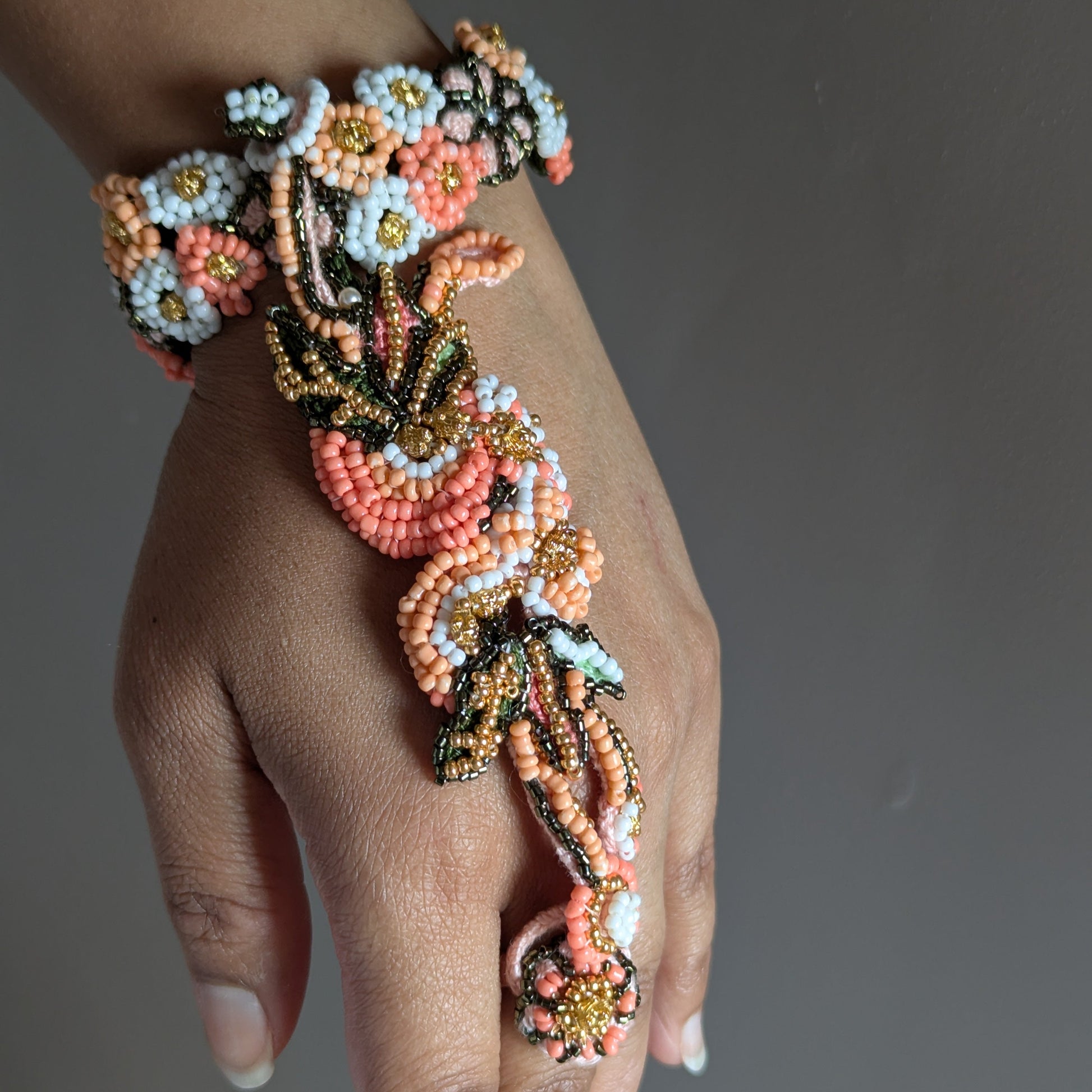A close-up of a hand wearing the one of a kind Padmika 5-in-1 Indian hand bracelet elaborate beaded in coral, peach, white, and gold seed beads with dark green accents, set against a solid dark grey background.