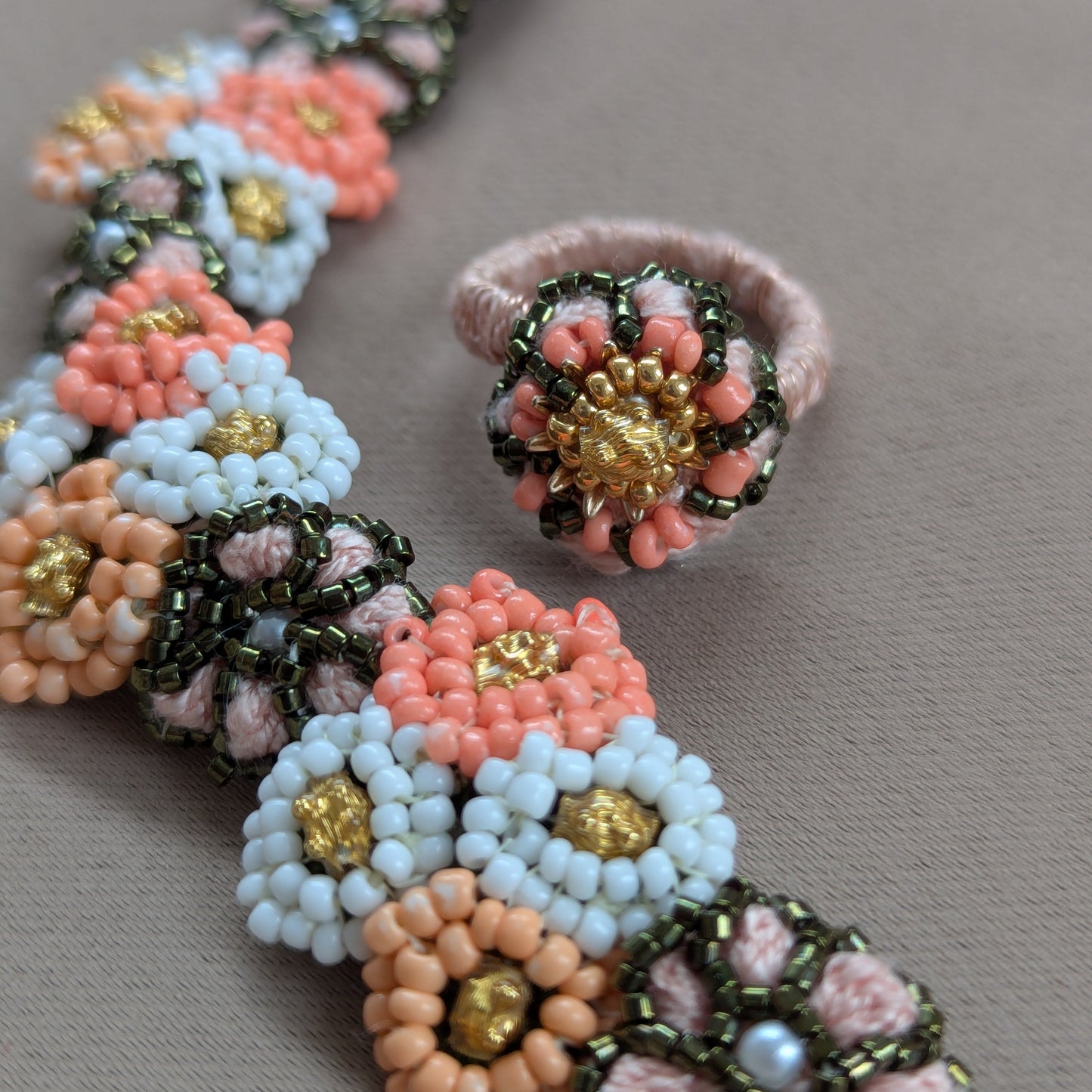 Close up view of the Padmika adjustable statement cocktail ring featuring peach, green and gold beads with the matching bracelet on a grey fabric backdrop.