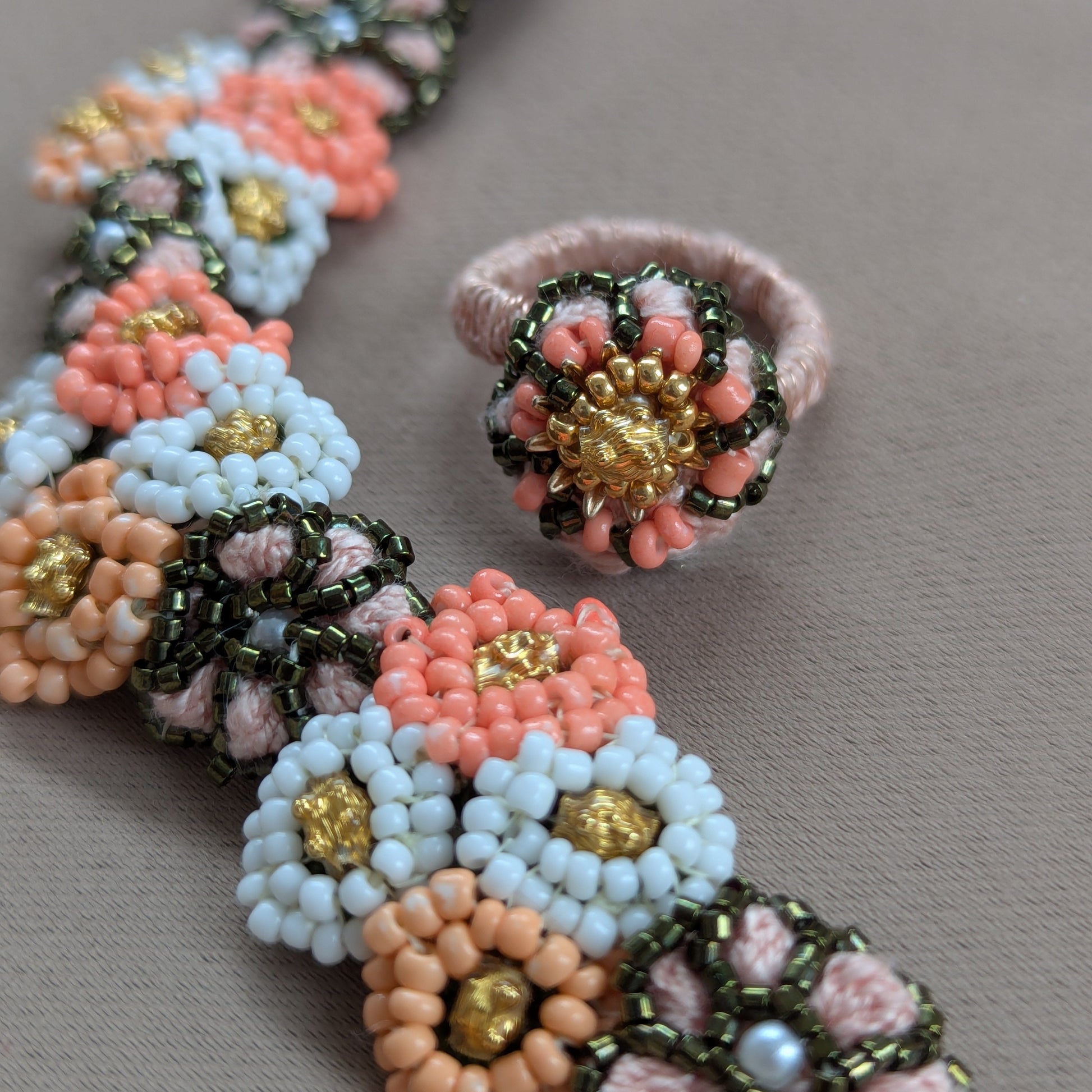 Close up view of the Padmika adjustable statement cocktail ring featuring peach, green and gold beads with the matching bracelet on a grey fabric backdrop.