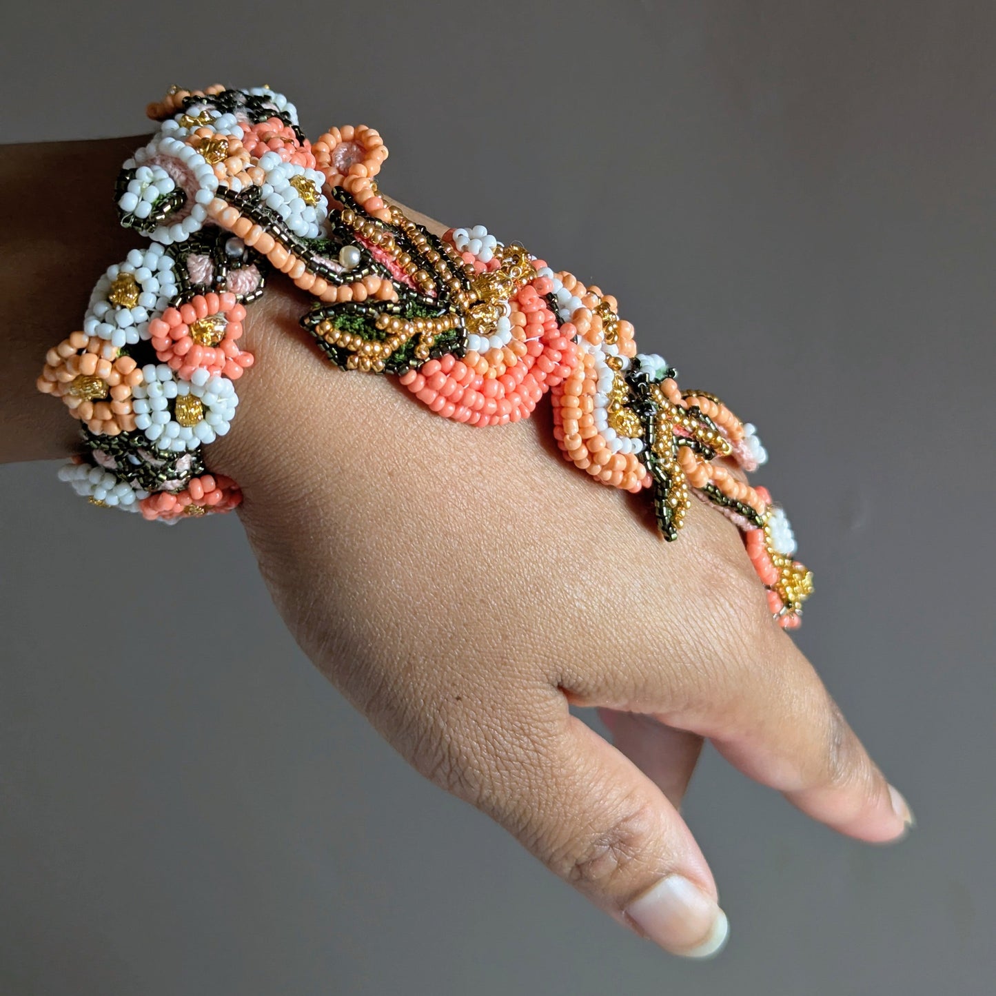 A close-up of a hand wearing the large Padmika 5-in-1 intricate beaded bracelet with floral and leaf patterns in coral, peach, white, black, and gold.