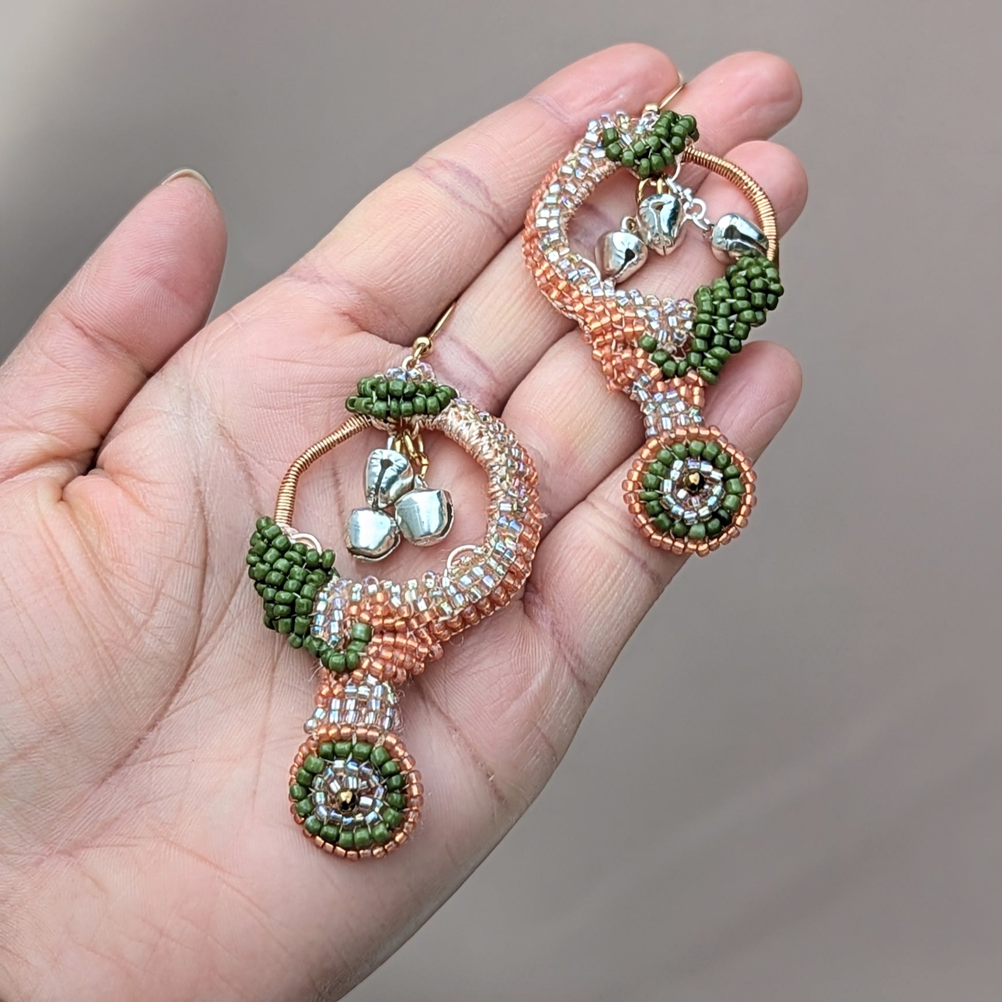 Hand holding a pair of one-of-a-kind Lakshmi beaded earrings featuring circular peach and emerald green patterns with dangling silver ghungroo bells.