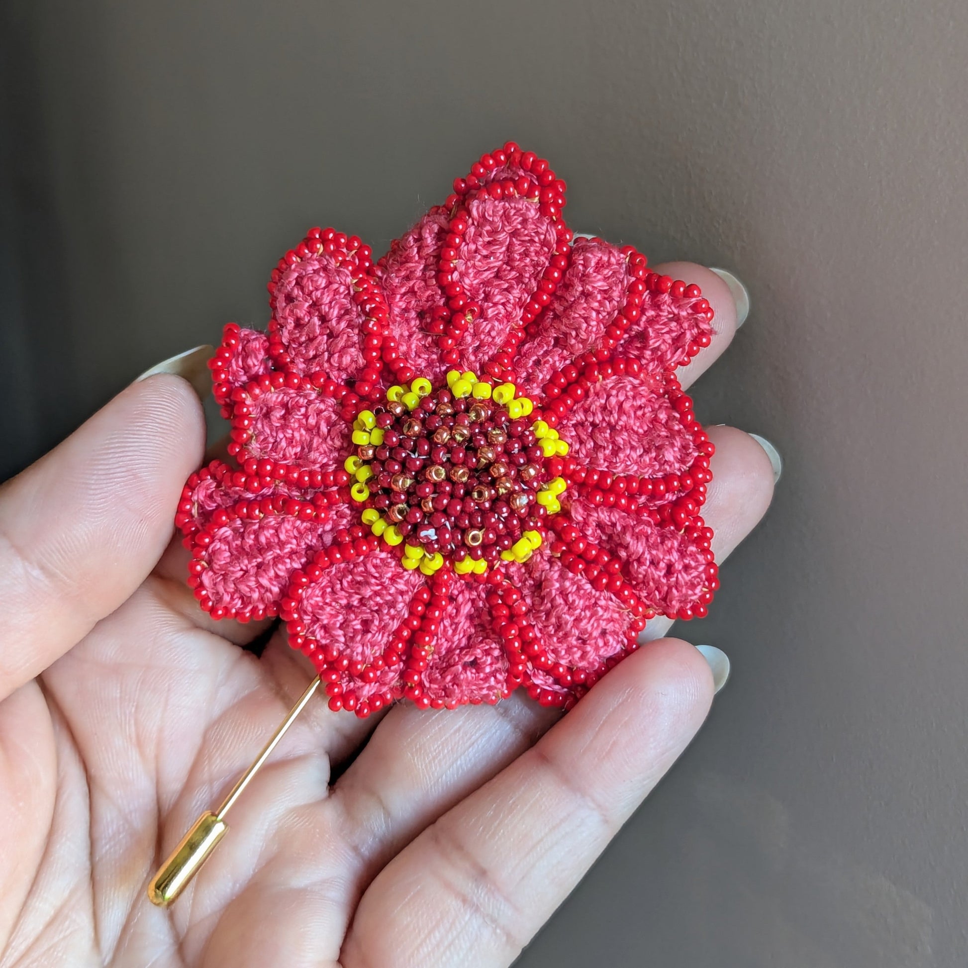 A one of a kind floral handmade Zinnia brooch pictured held in hand against a soft grey backdrop featuring hand crochet and hand beadwork artistry.