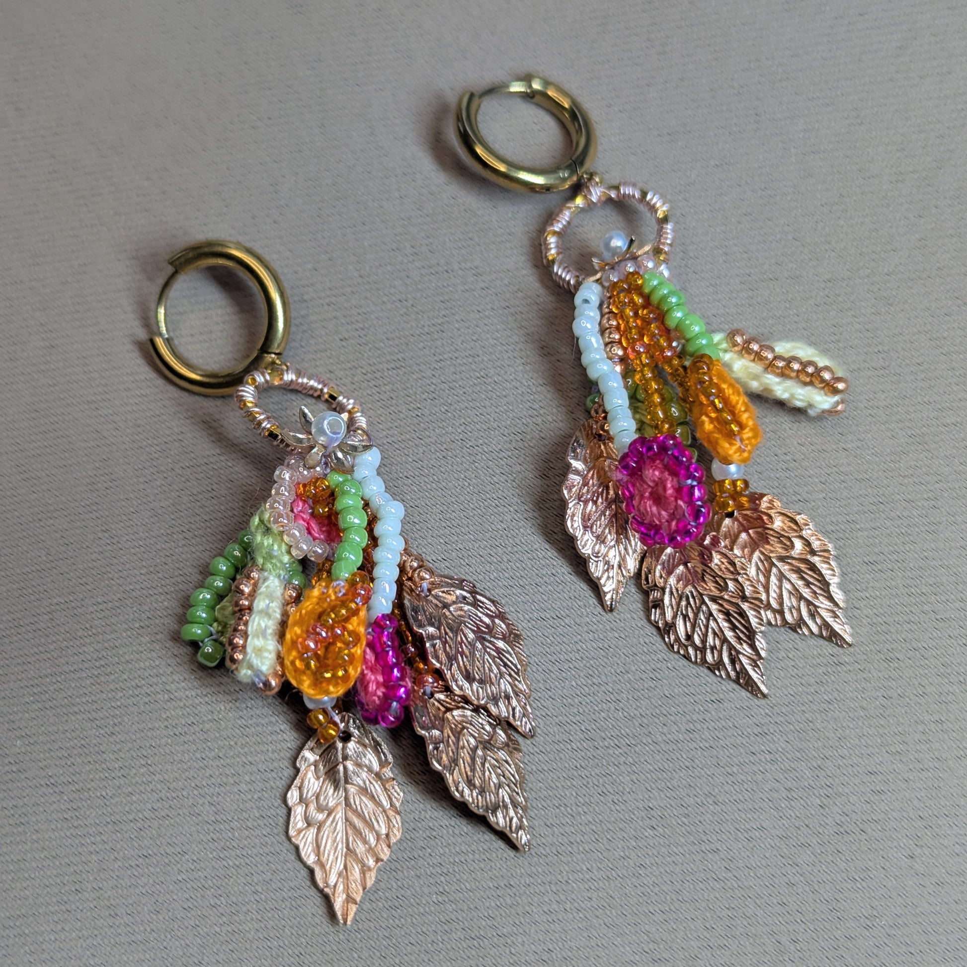 Flat lay of Zariya one-of-a-kind spring garden earrings with rose gold plated leaf charms, seed pearls and pastel beaded strands on a neutral fabric background.