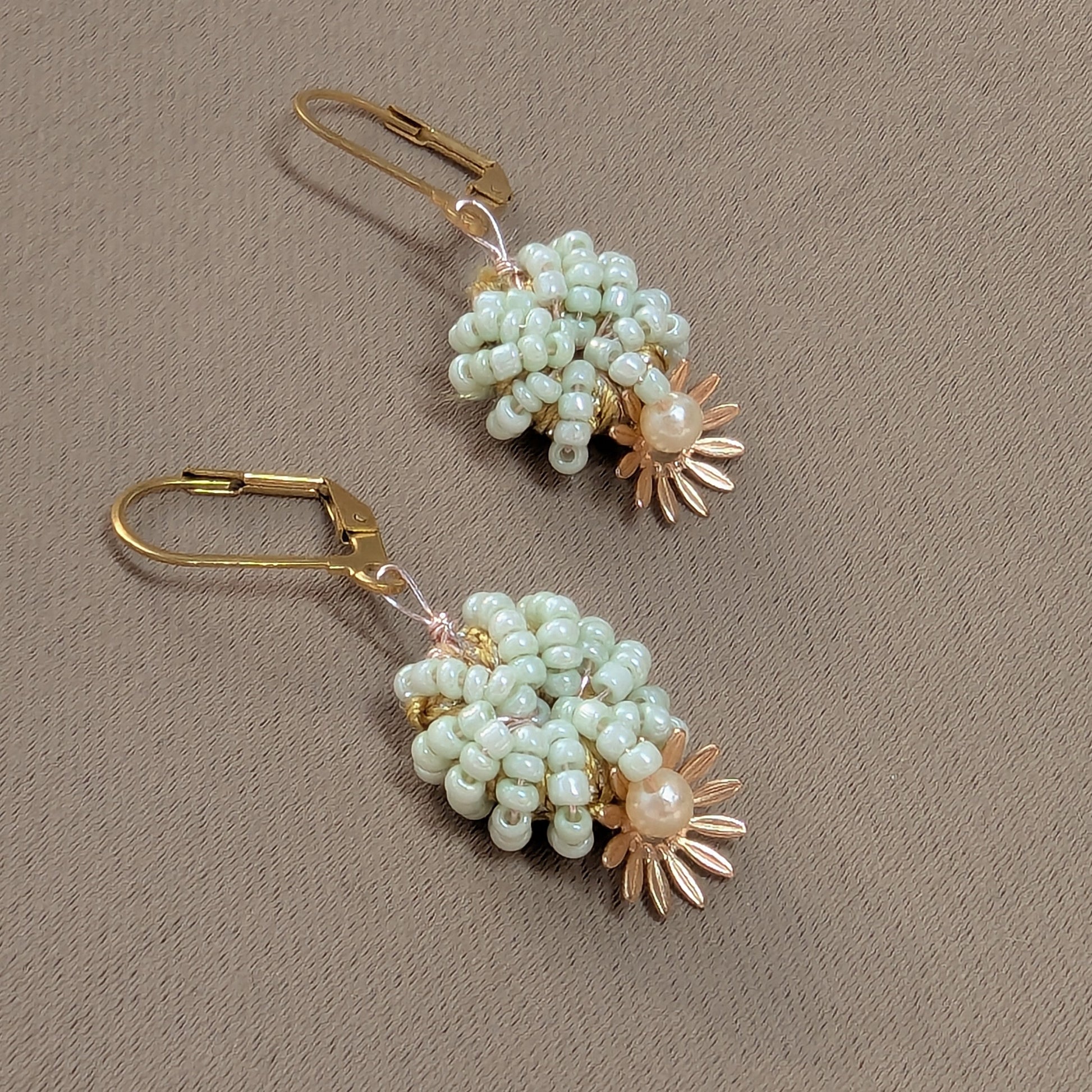 A pair of beaded gold and mint green handcrafted earrings against a soft neutral grey fabric backdrop.