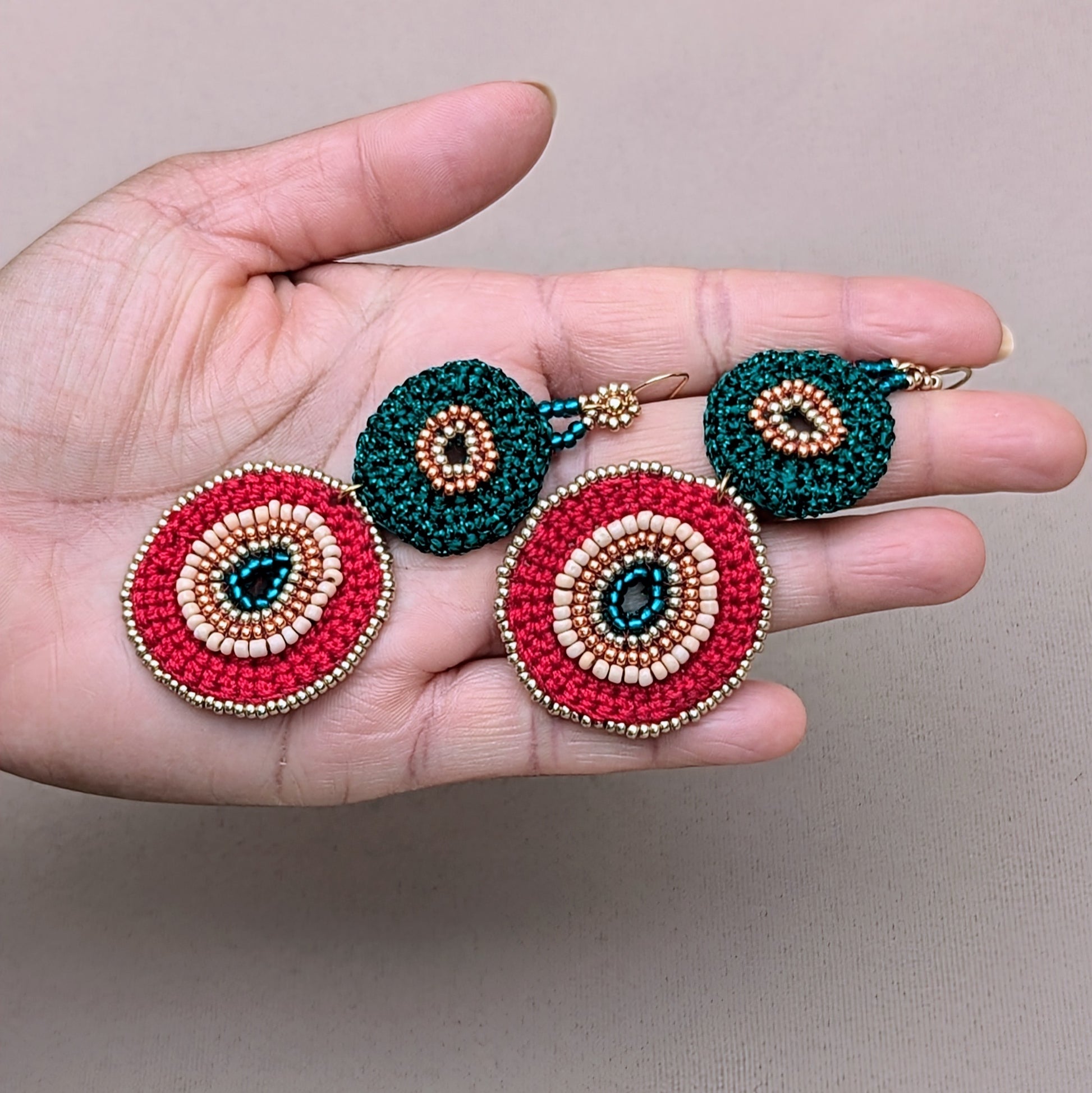 Hand holding a large statement pair of green, red, and gold circular element earrings handcrafted in a mixture of hand beadwork and hand crochet.
