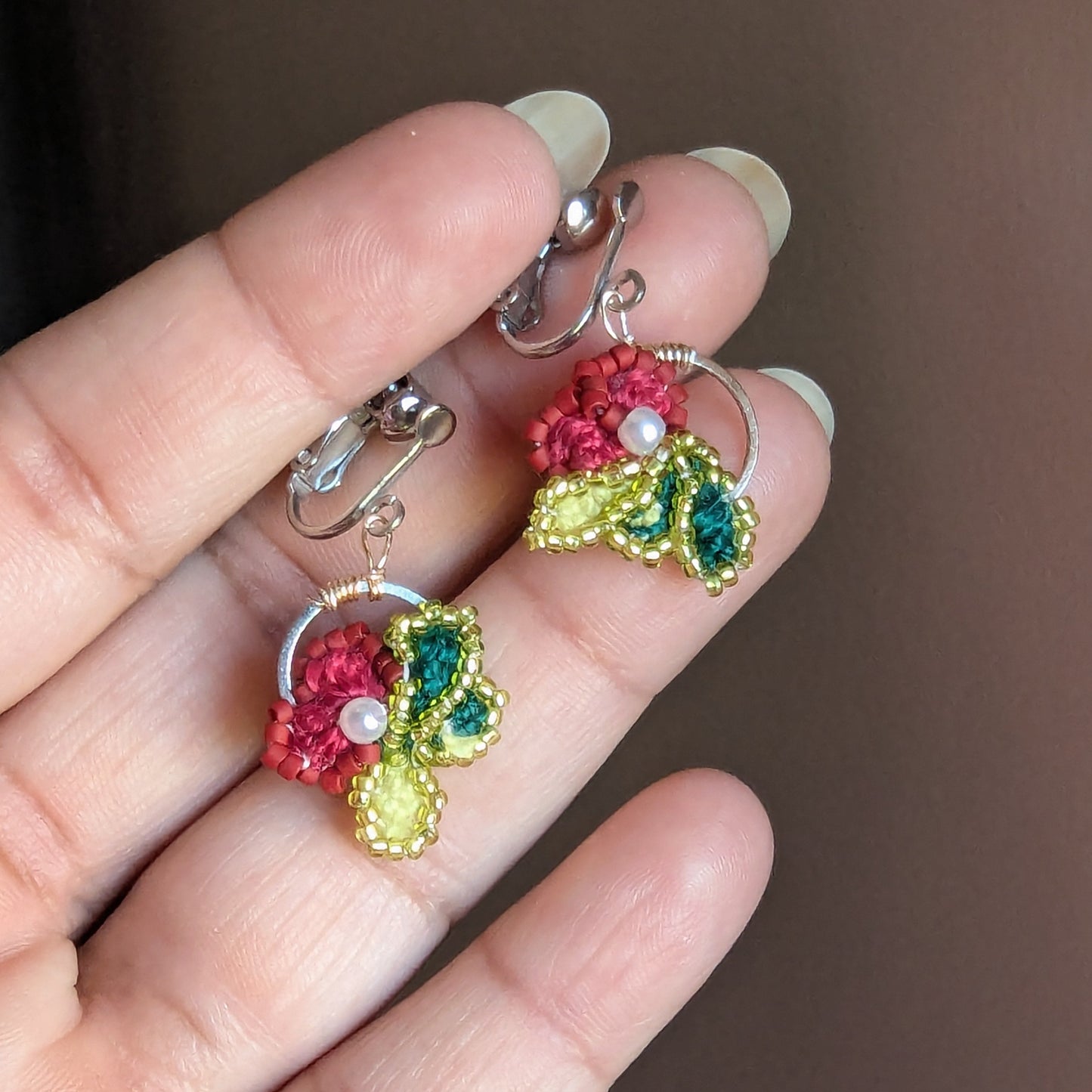 Reva clip-on earrings held between fingertip showing stainless steel screw-back clasp and hoop frame.