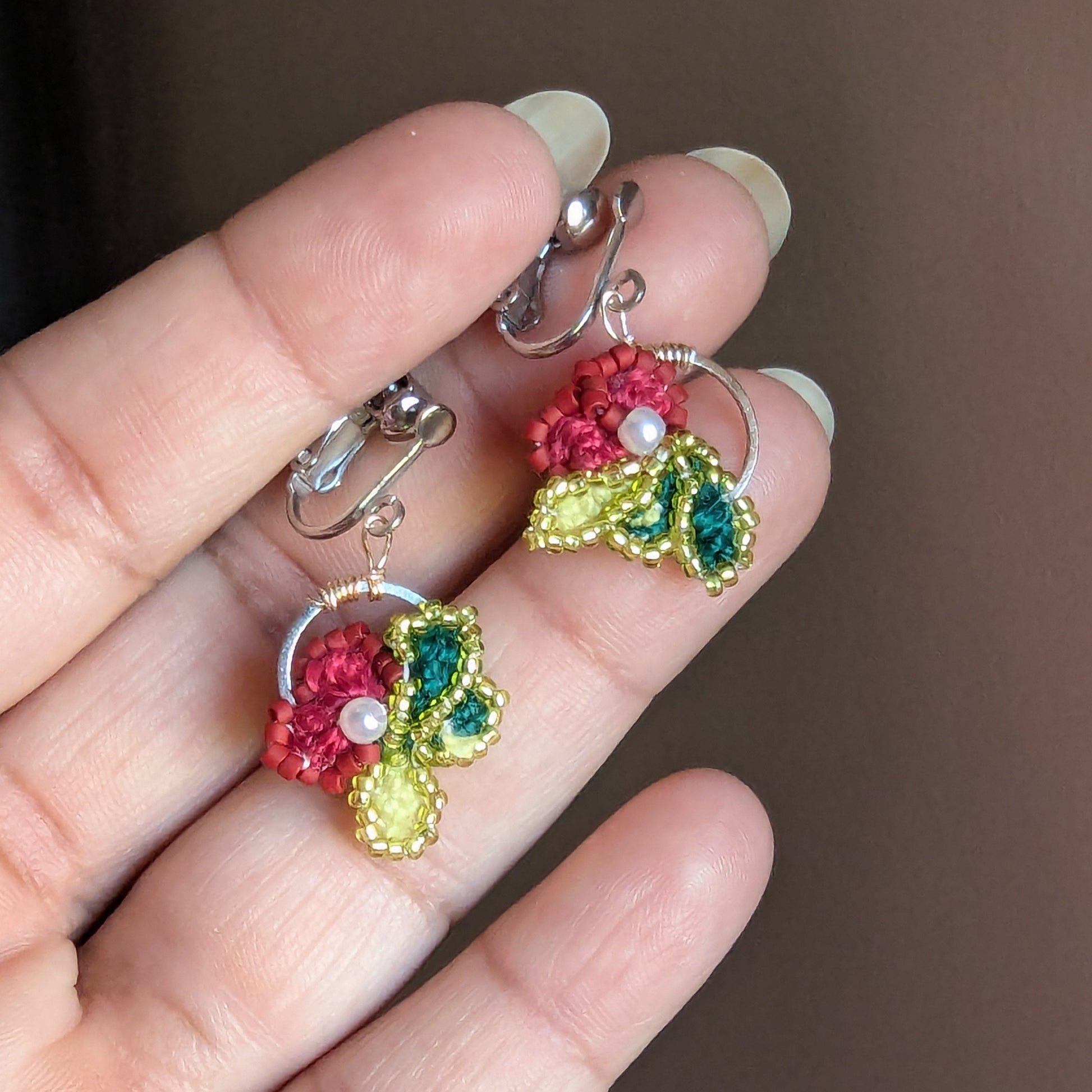 Reva clip-on earrings held between fingertip showing stainless steel screw-back clasp and hoop frame.