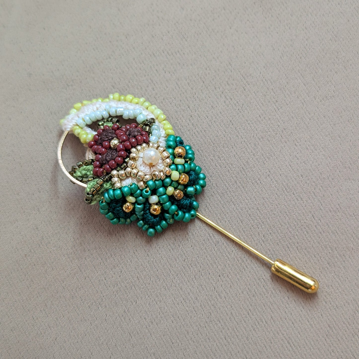 Sarika one of a kind brooch held in hand with layered green beadwork and gold stainless steel pin pictured against a neutral fabric backdrop.
