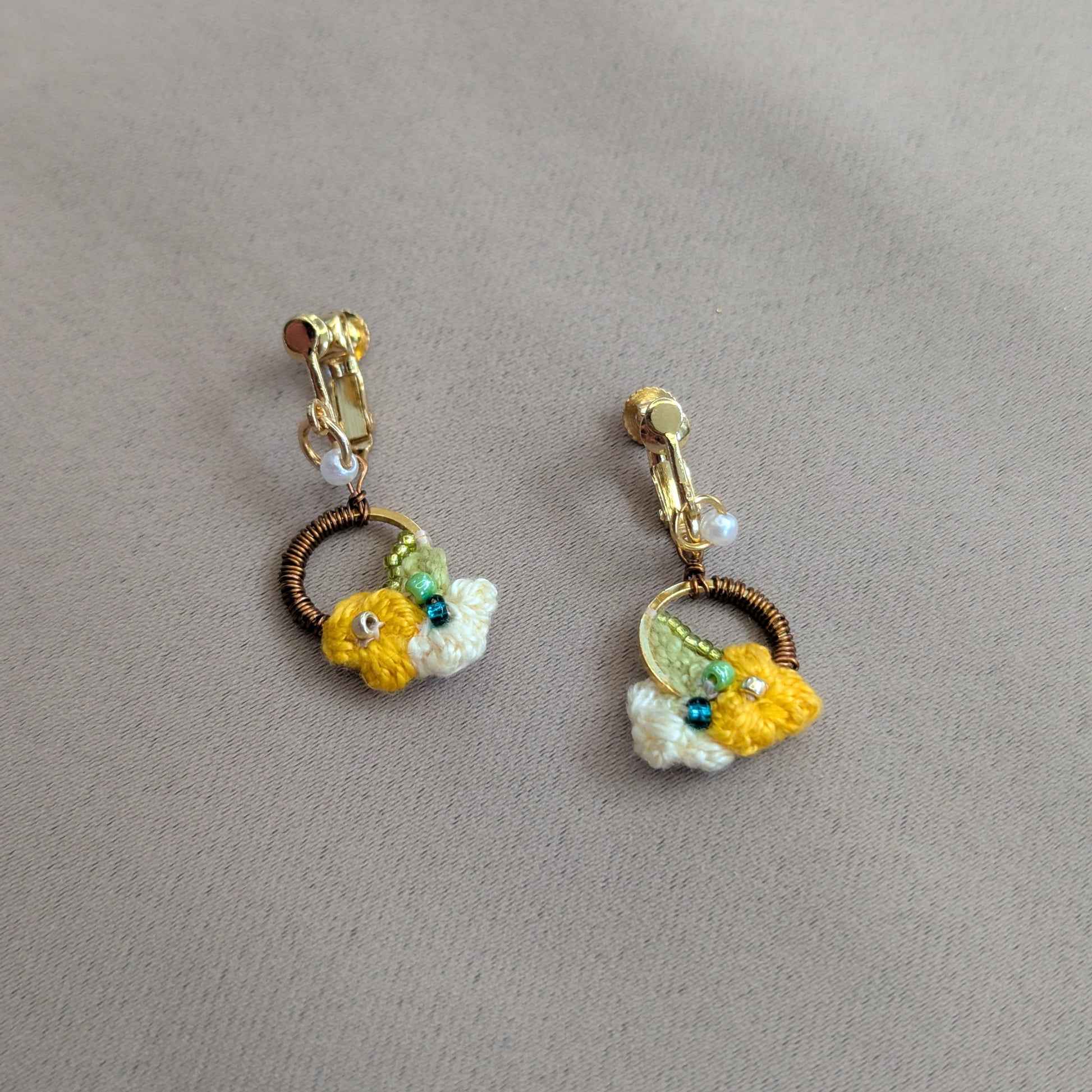 A pair of Sauri handmade gold stainless hoop clip-on earrings with yellow and cream floral motifs, green, gold, bronze and pearl accents exclusively available at Mayaani Jewellery UK.