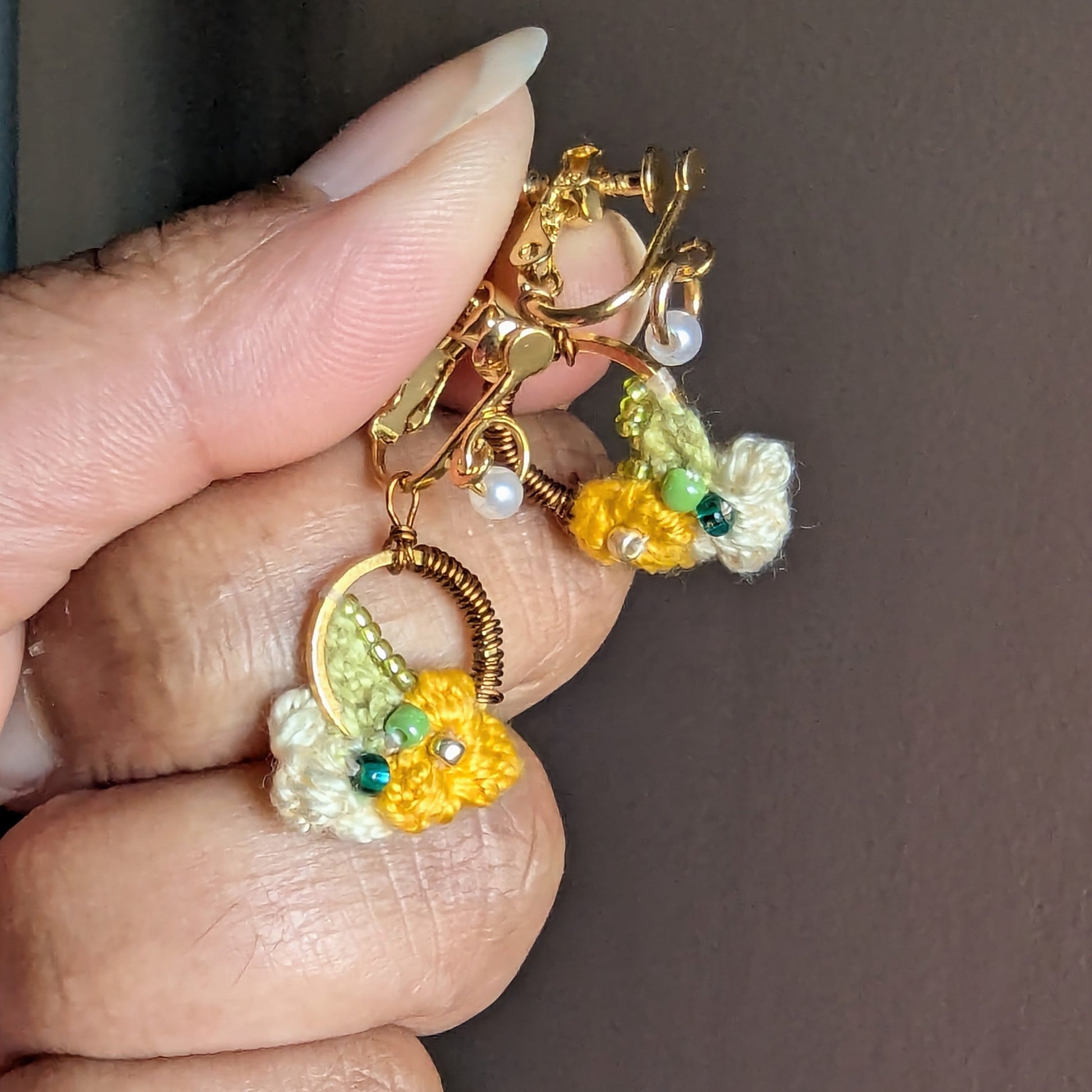 Hand holding a delicate artisan made pair of clip-on Sauri earrings in yellow and cream florals with gold, bronze, pearl and bronze accents in seed beads, wire artistry and crochet for comfortable heirloom quality style for non-pierced ears.